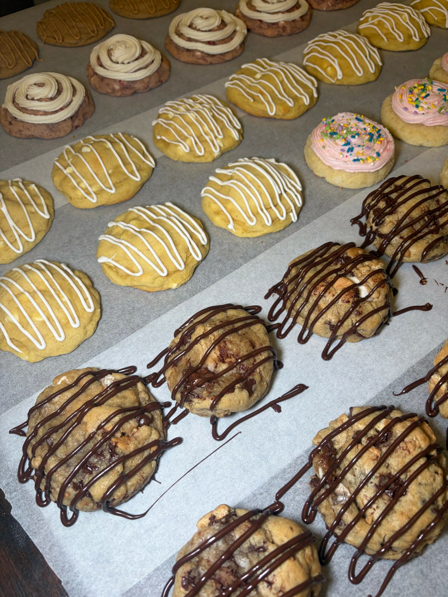 Assorted cookies with icing and sprinkles on parchment paper, some drizzled with chocolate, arranged in rows.