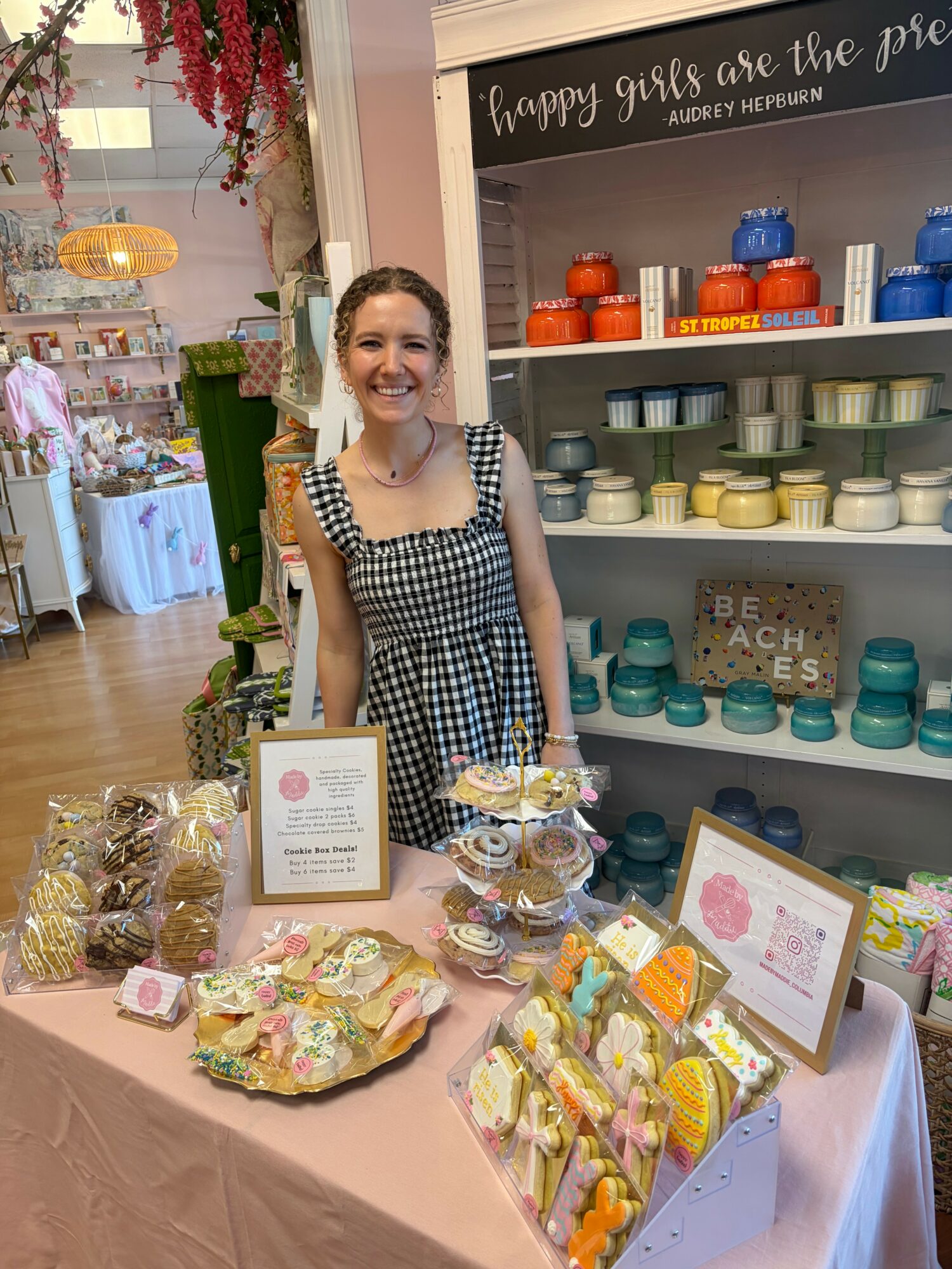 Young woman standing behind a table with colorful baked goods and display signs, in a store or market setting.