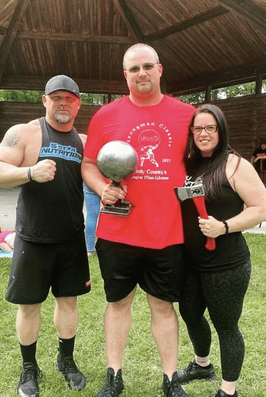 Three people standing outdoors under a gazebo, holding a trophy and drinks, smiling at the camera.