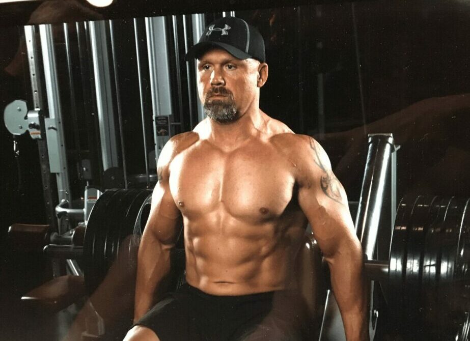 Muscular man with a beard and cap in a gym, shirtless, sitting on a workout bench.