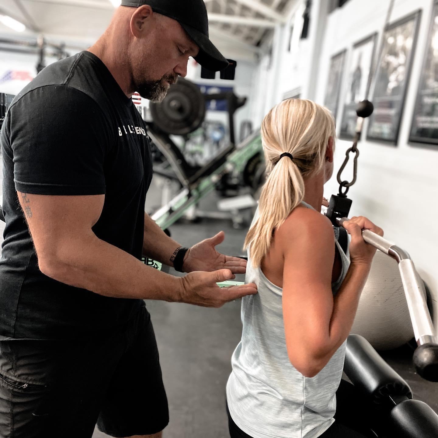 Personal trainer assisting a woman with a cable exercise in a gym, with framed photos on the wall.