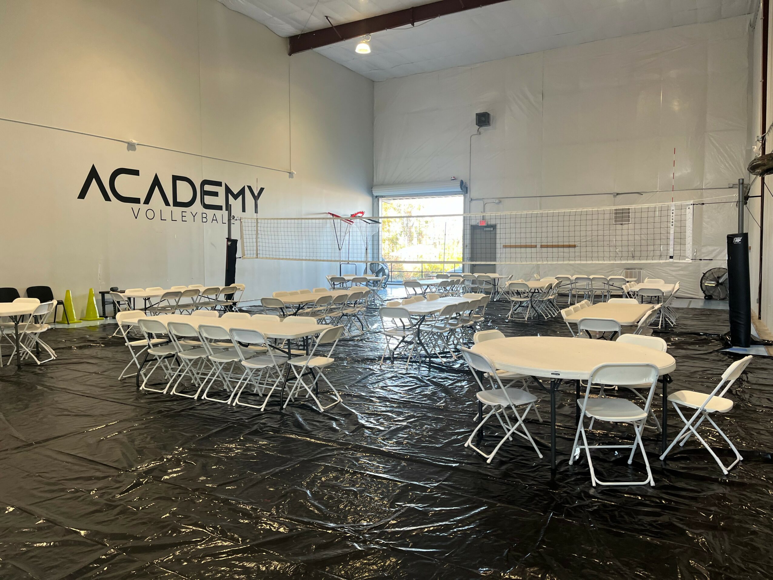 Indoor volleyball court with tables and chairs set up, wall with 'ACADEMY VOLLEYBALL' sign, net, and window.