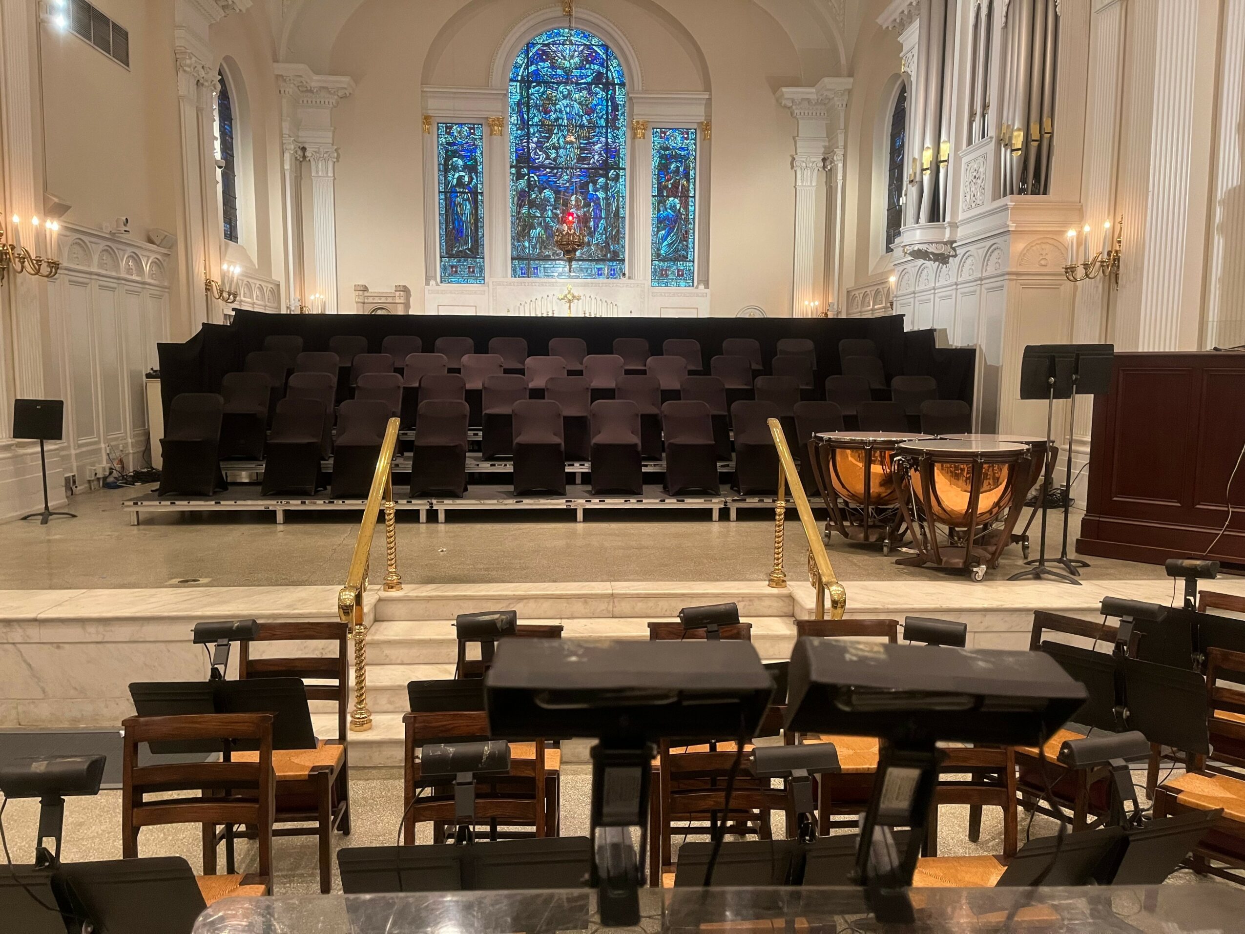 Interior of a church with a stage, chairs, and musical instruments, stained glass window at the back, and ornate architecture.