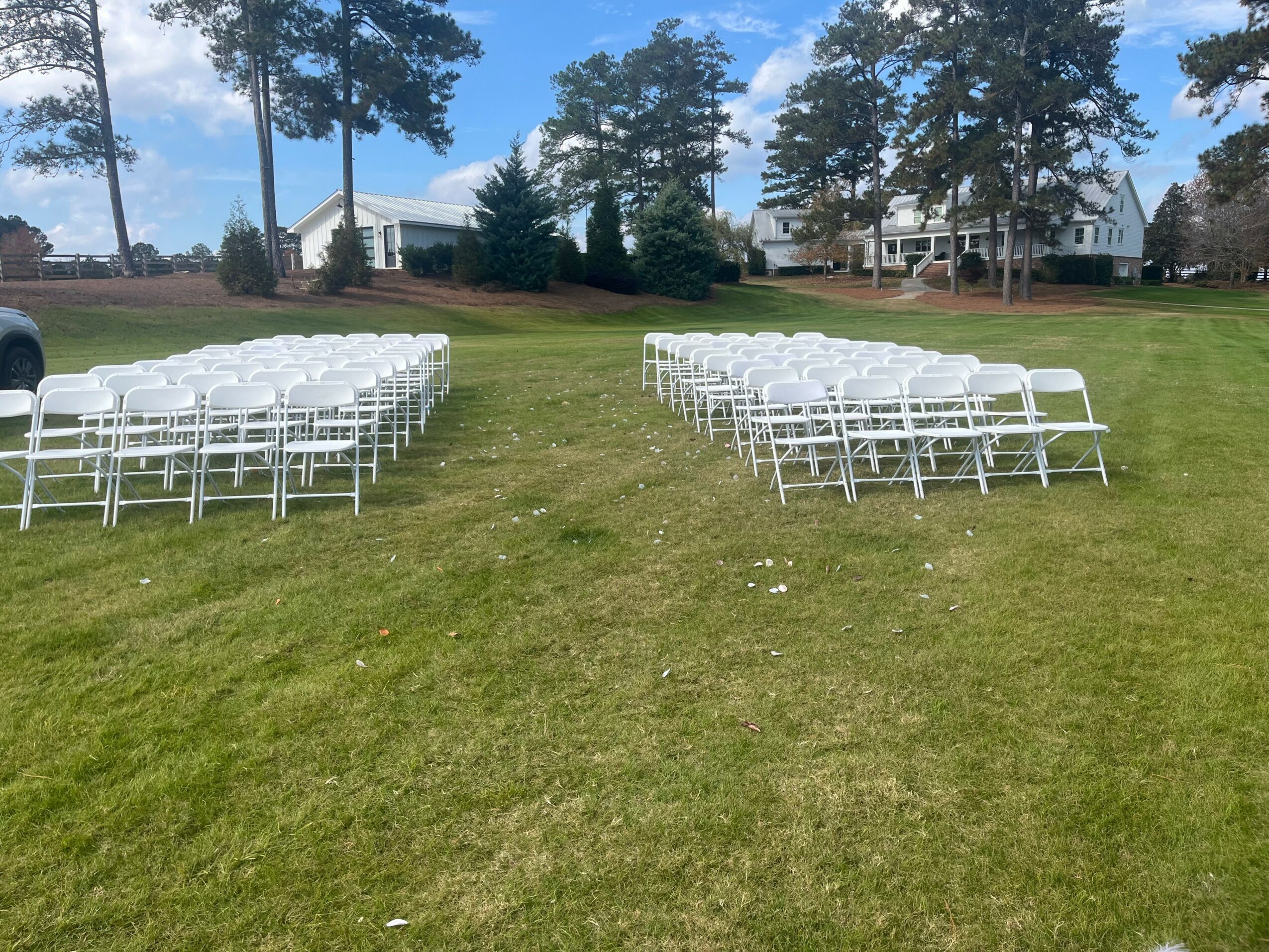 Rows of white chairs set up on a grassy field with trees and houses in the background.