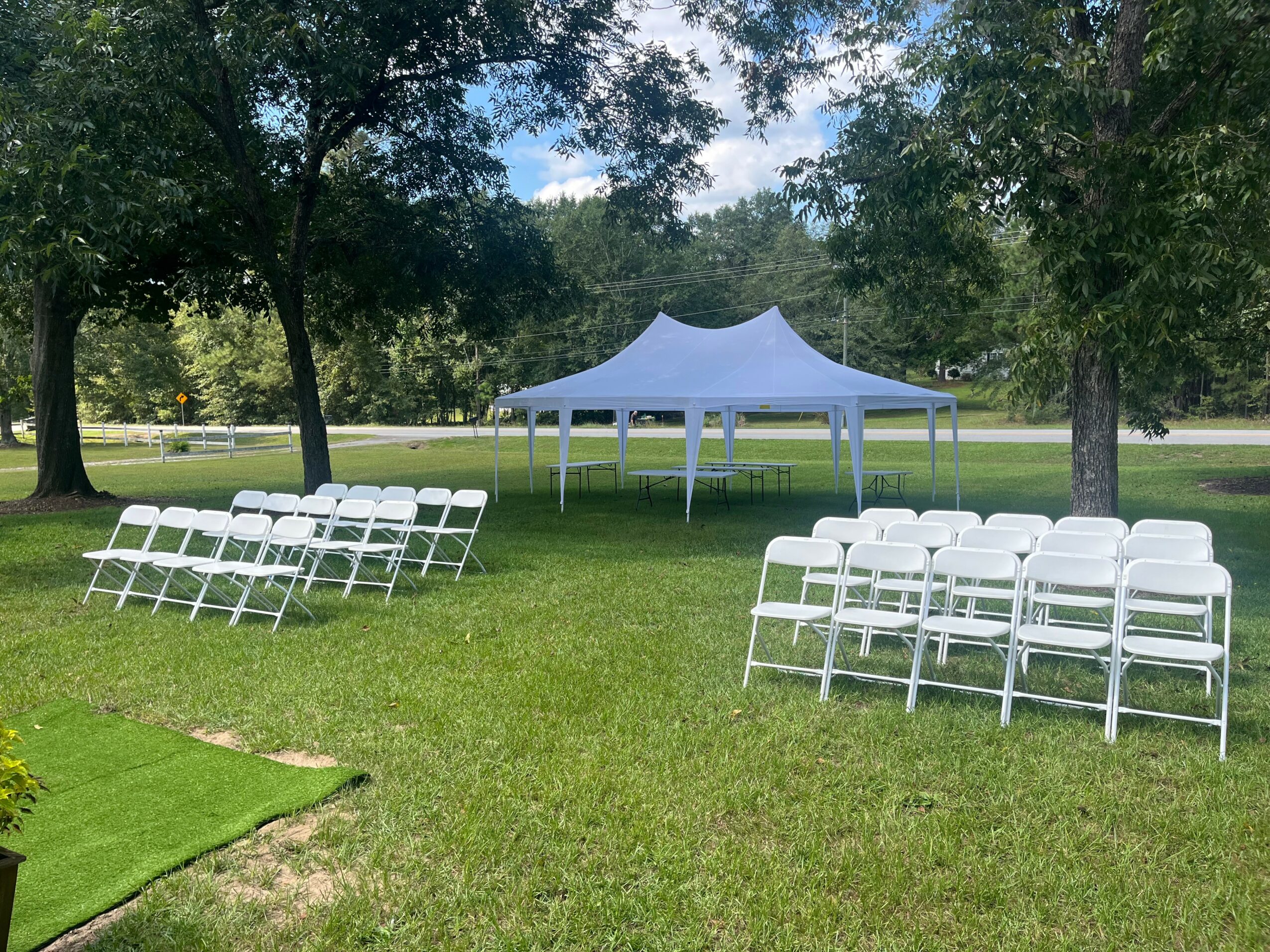Outdoor setup with white chairs arranged in rows on grass, white canopy tent in background, trees and blue sky.