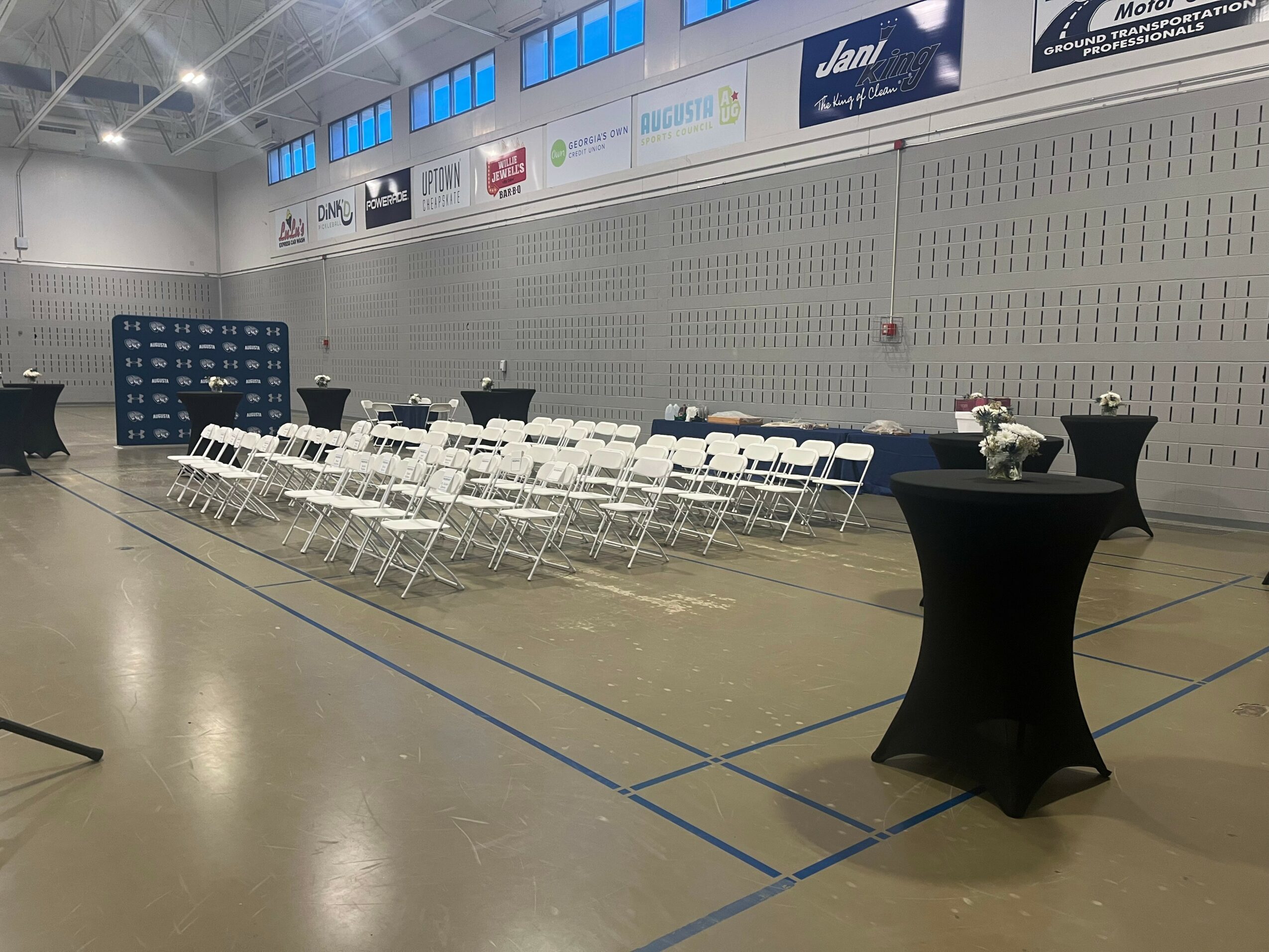 Event setup with rows of white chairs and black cocktail tables in a gymnasium.