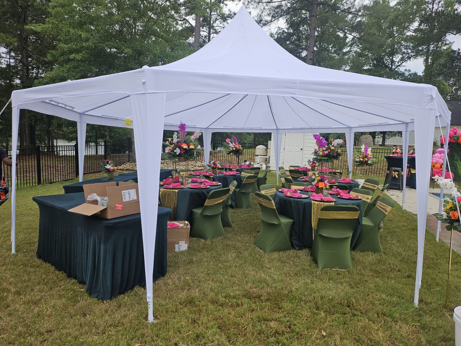 Outdoor event setup with round tables, green chairs, pink table settings, and a white canopy tent on grass.