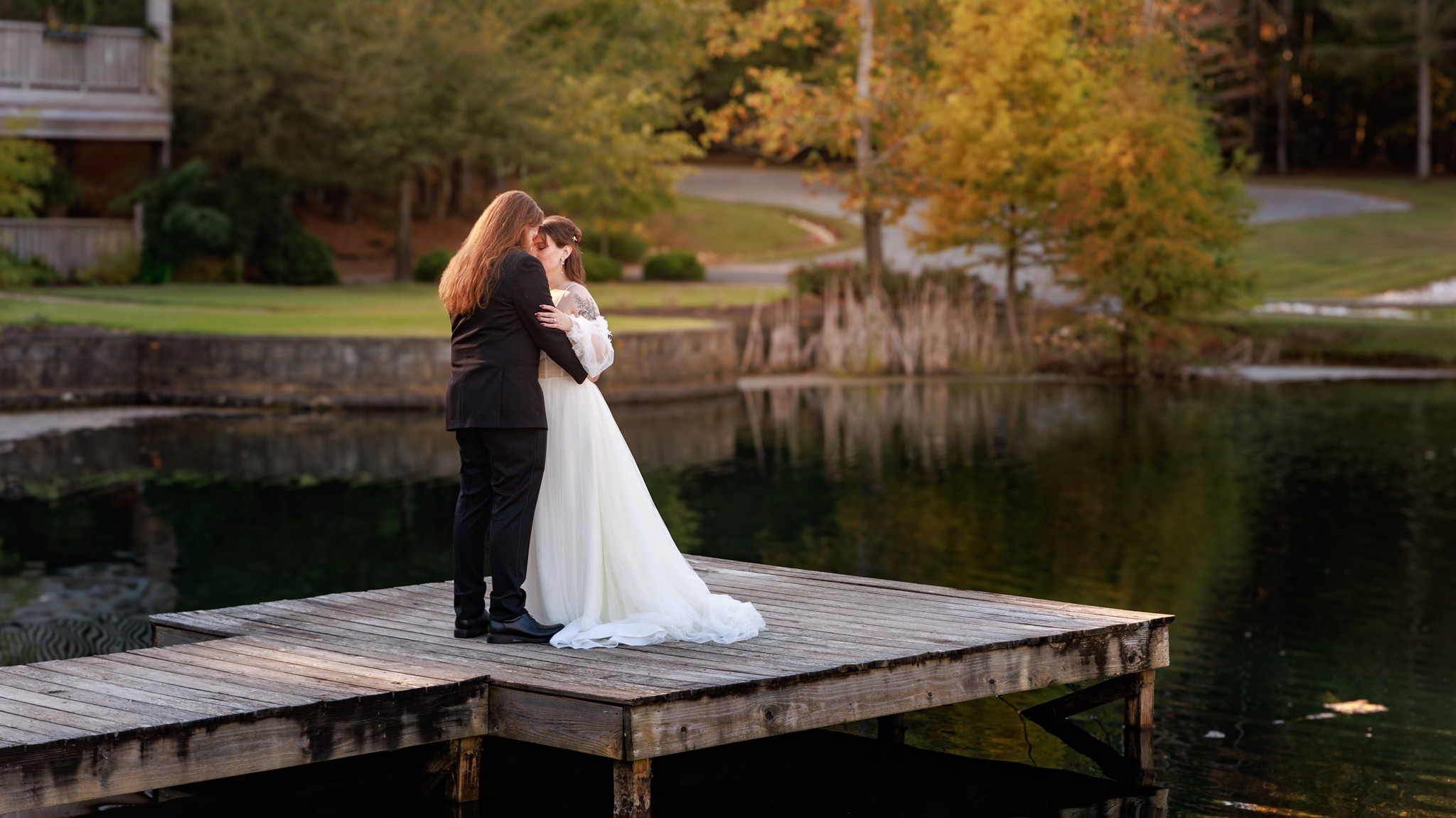 Two women in wedding attire embrace on a wooden dock by a lake with trees and a house in the background.