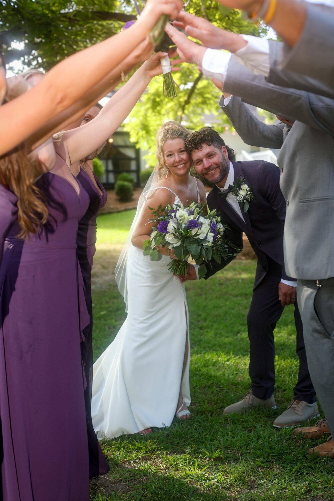 Bride and groom smiling, holding a bouquet, surrounded by friends raising hands outdoors with trees and grass.