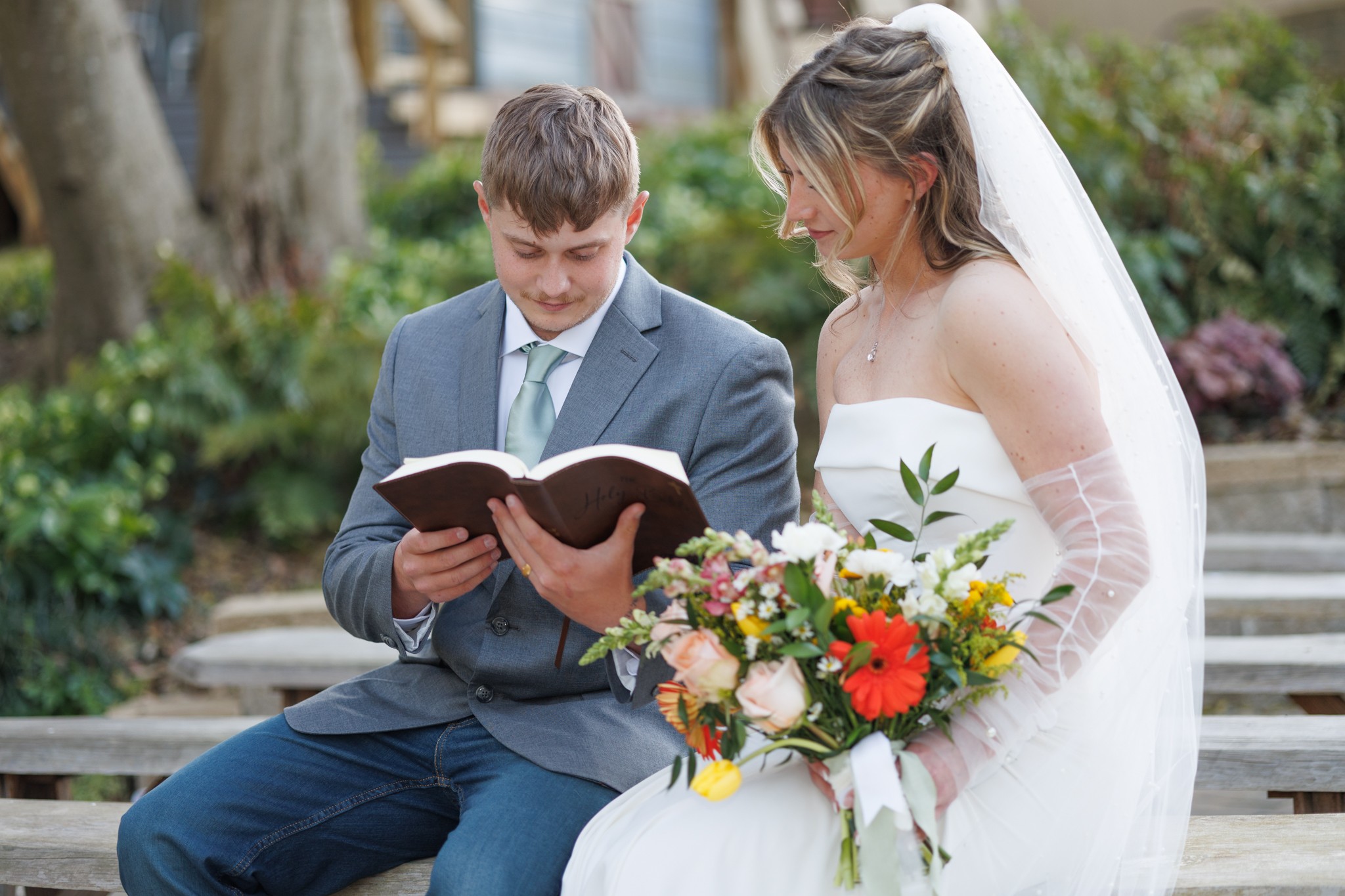 Bride and groom sit on a bench outdoors, reading a book together, with greenery in the background.