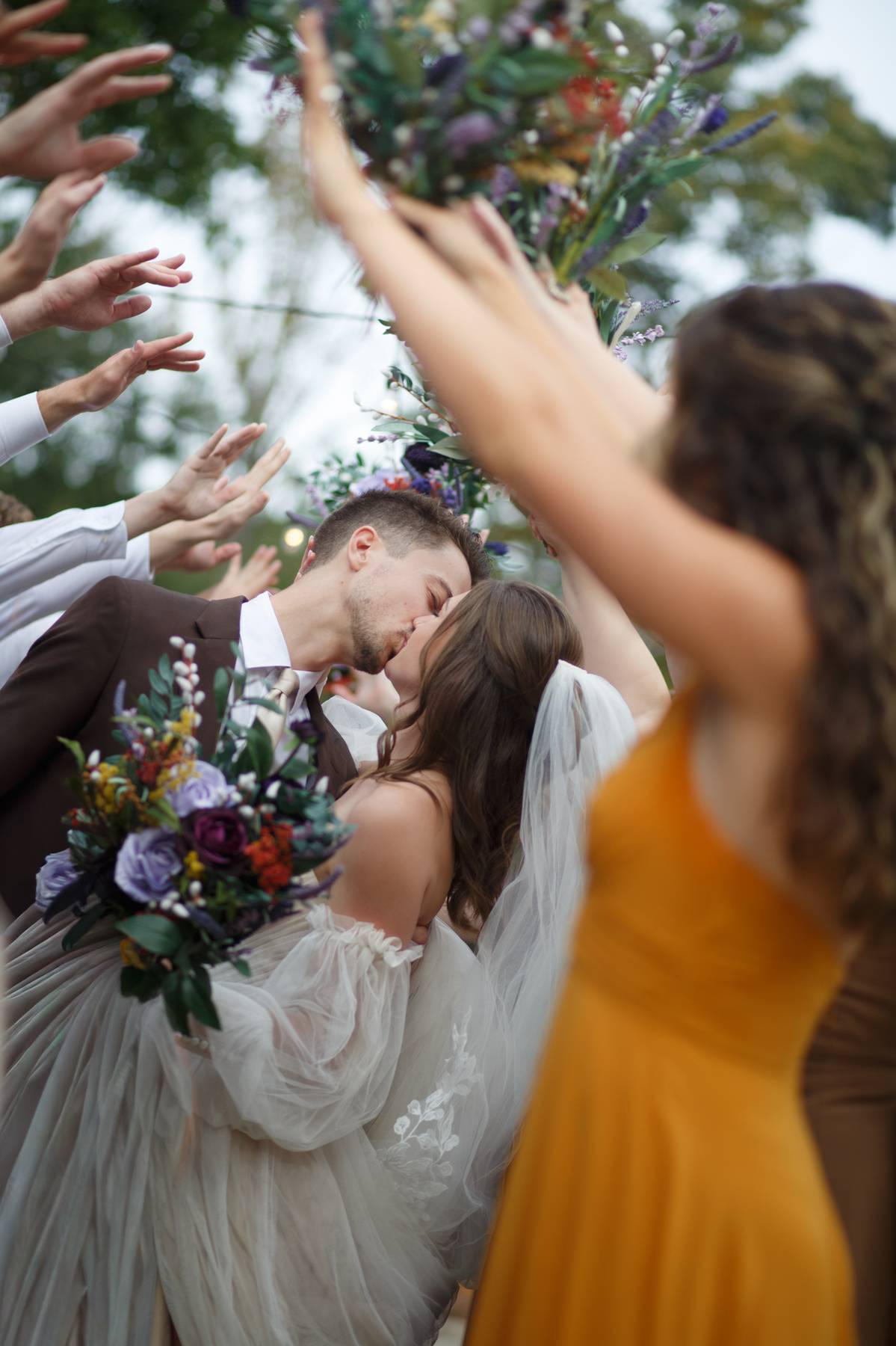 Bride and groom kissing surrounded by people throwing flowers outdoors.