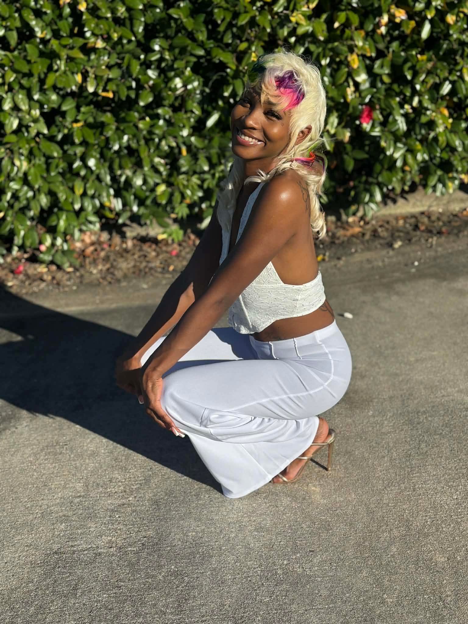 Woman crouching on pavement, smiling, with pink hair and white outfit, greenery in background.