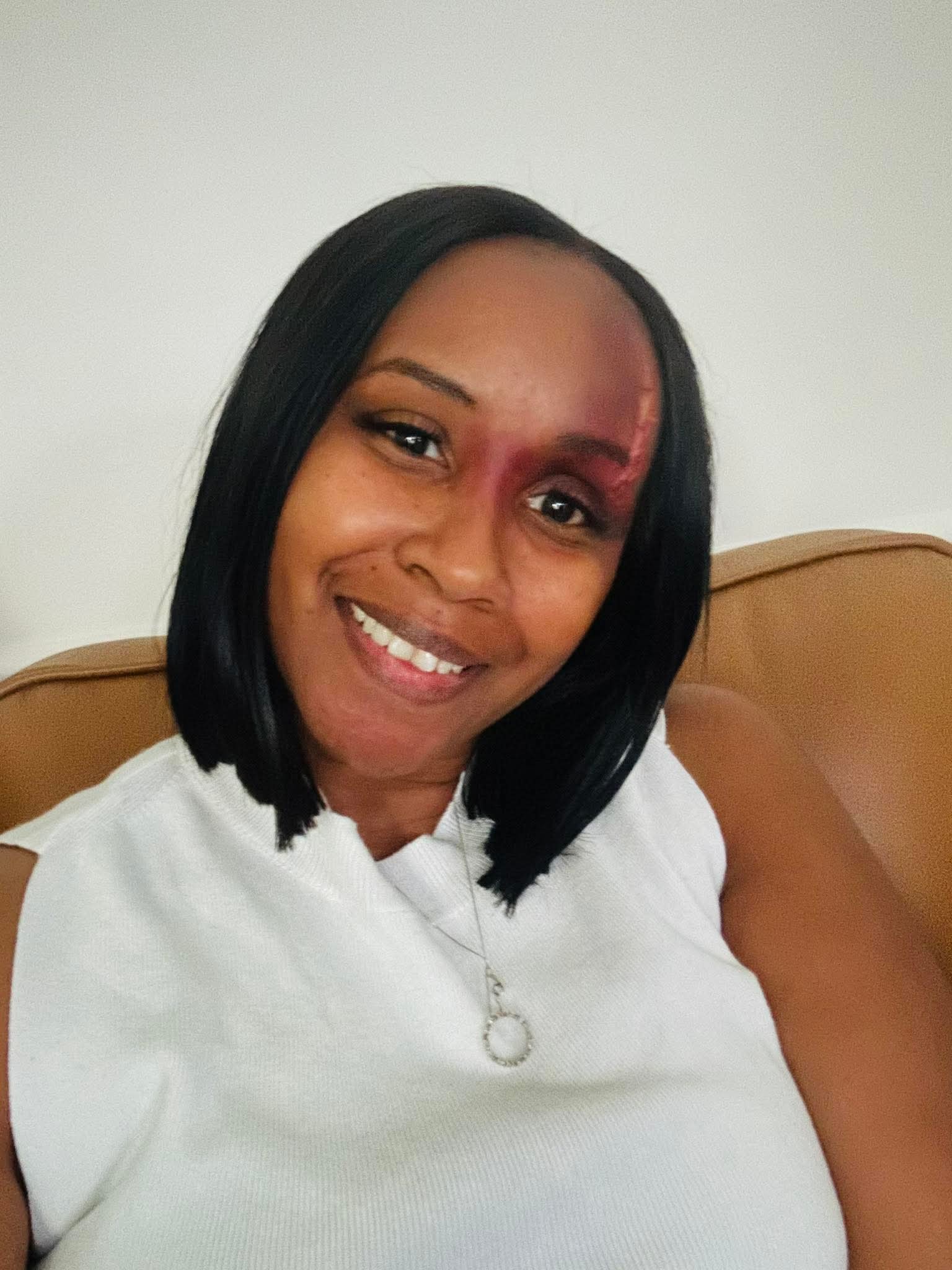 Woman with shoulder-length black hair smiling, wearing a white sleeveless top, sitting on a beige couch.