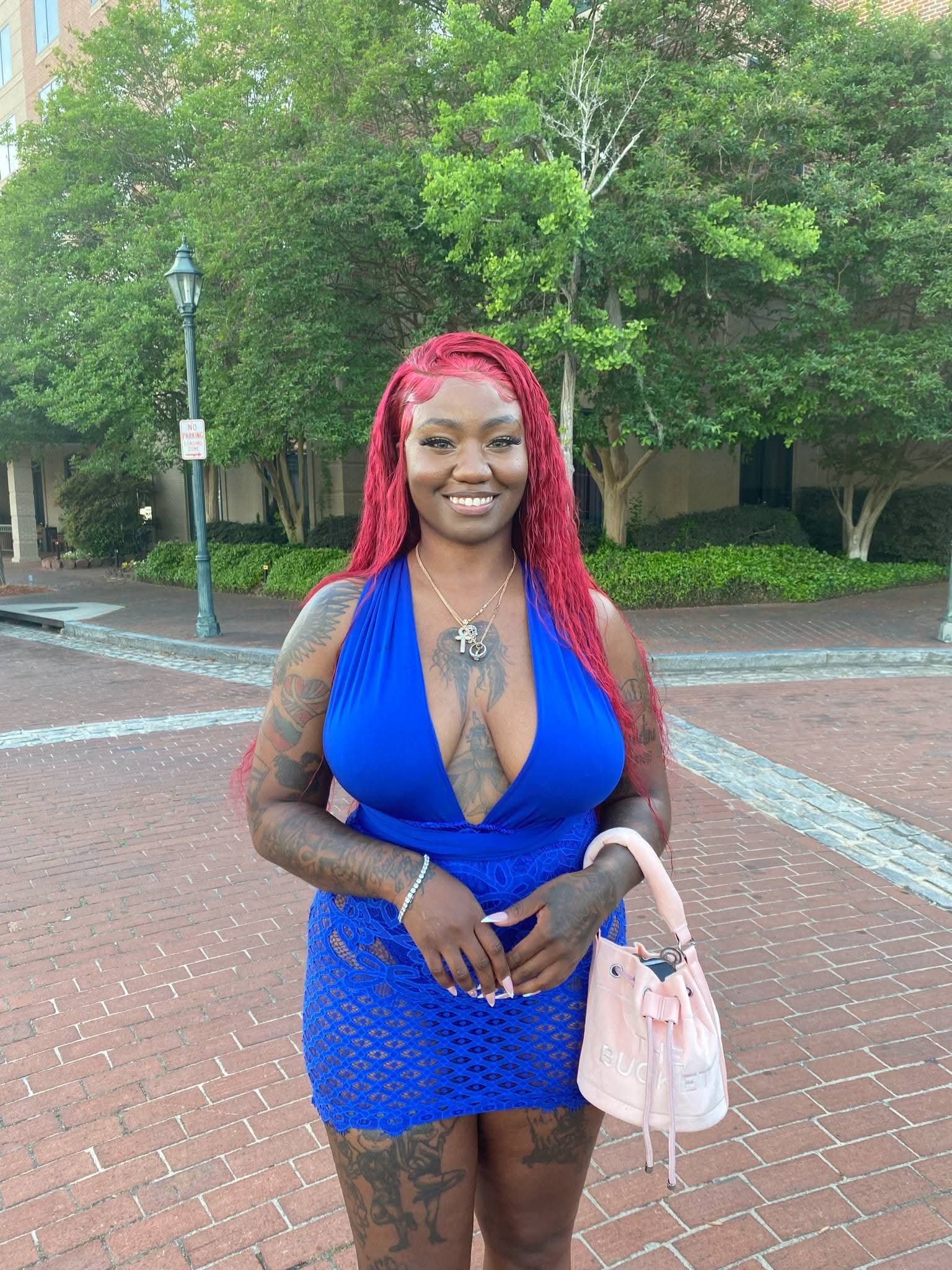 Woman with red hair in a blue dress standing outdoors on brick pavement, holding a pink bag, smiling.