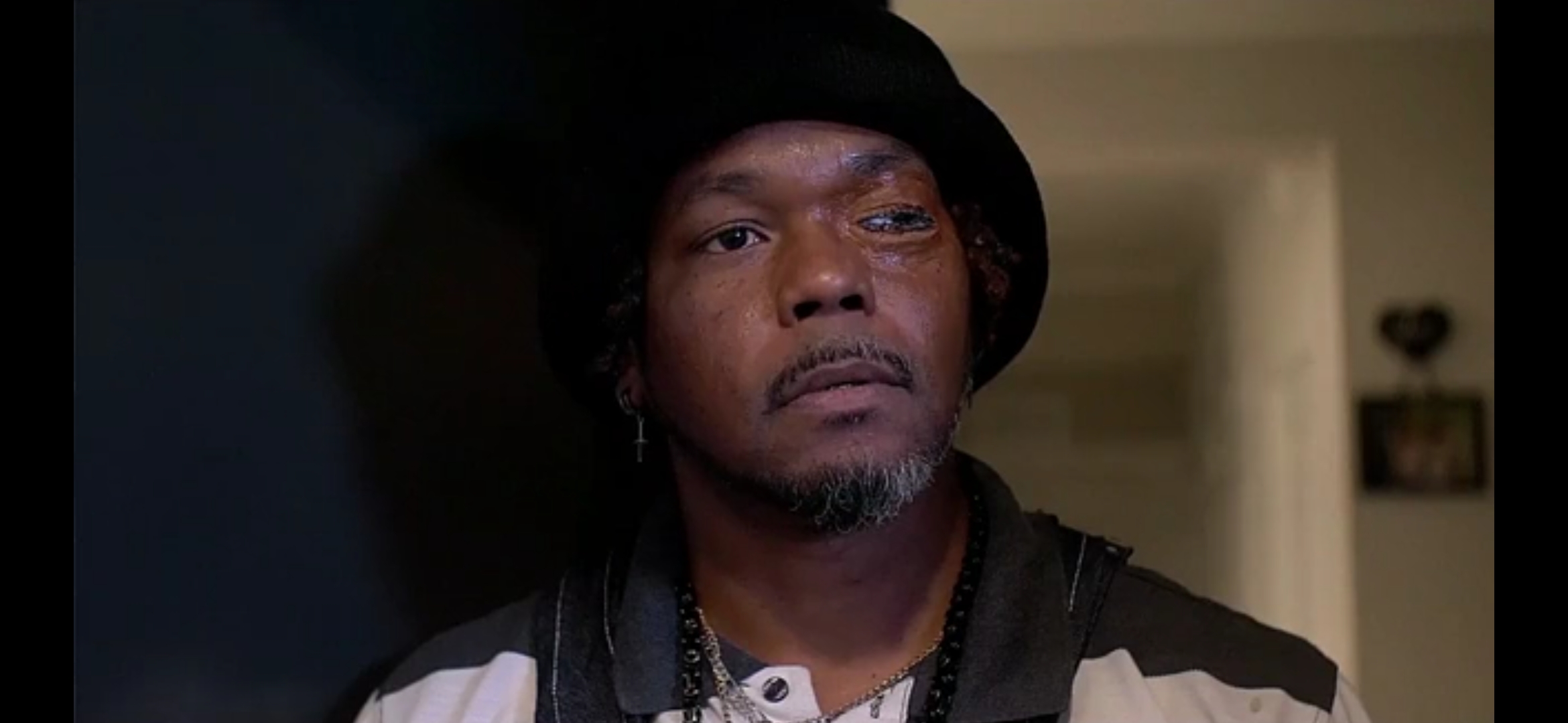 Man with dark skin, wearing a black hat and dark clothing, looking serious indoors.
