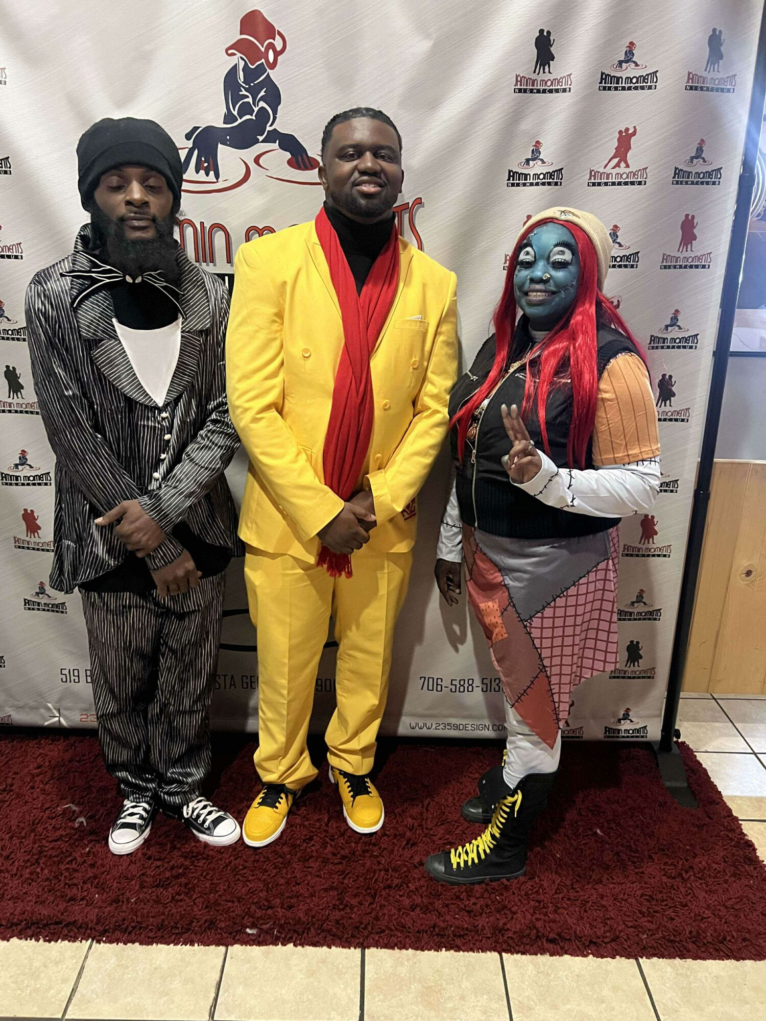 Three people standing on a red carpet in front of a backdrop with logos, smiling and posing for the photo.