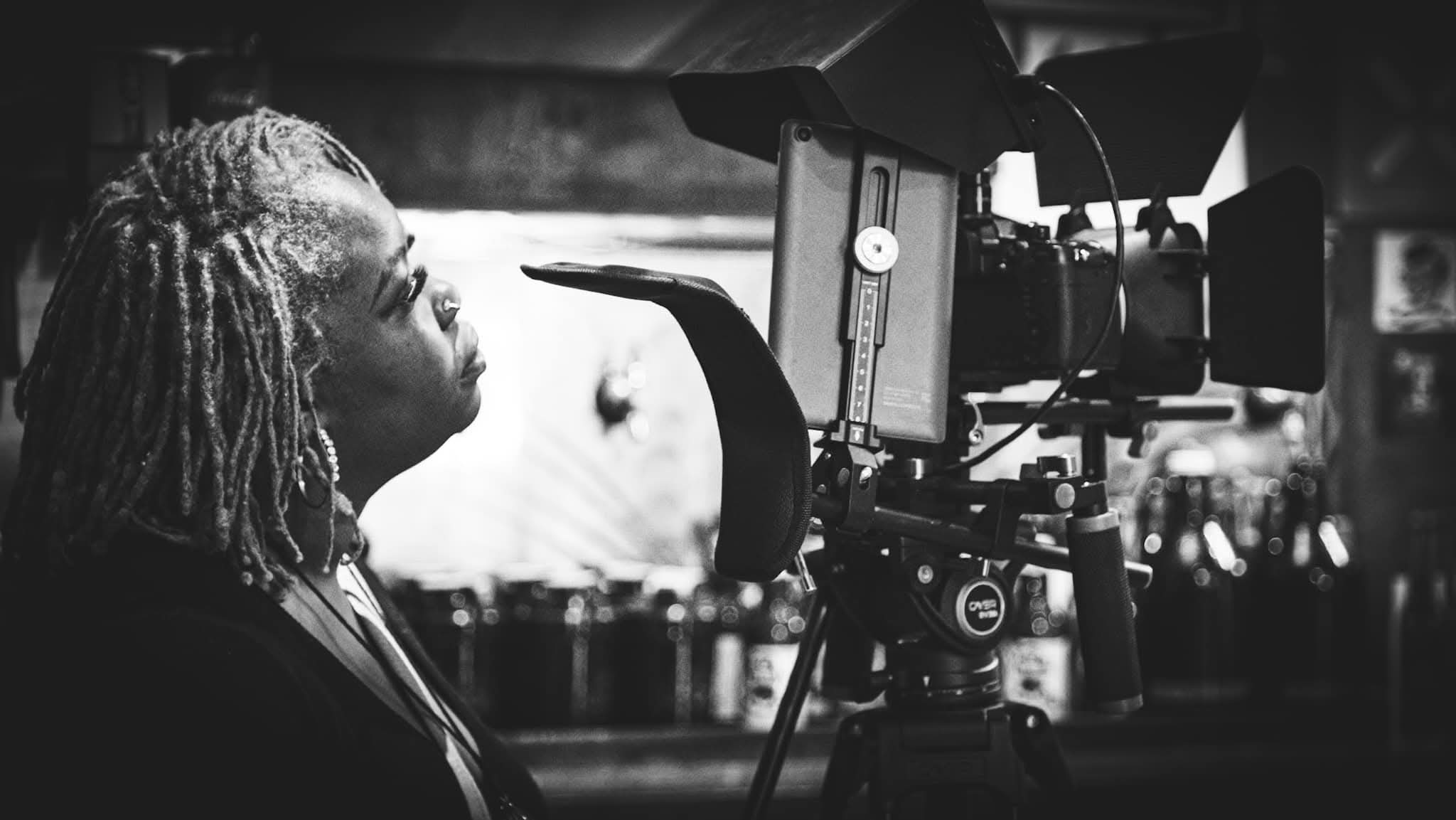 Person with dreadlocks operating a film camera indoors, background blurred.