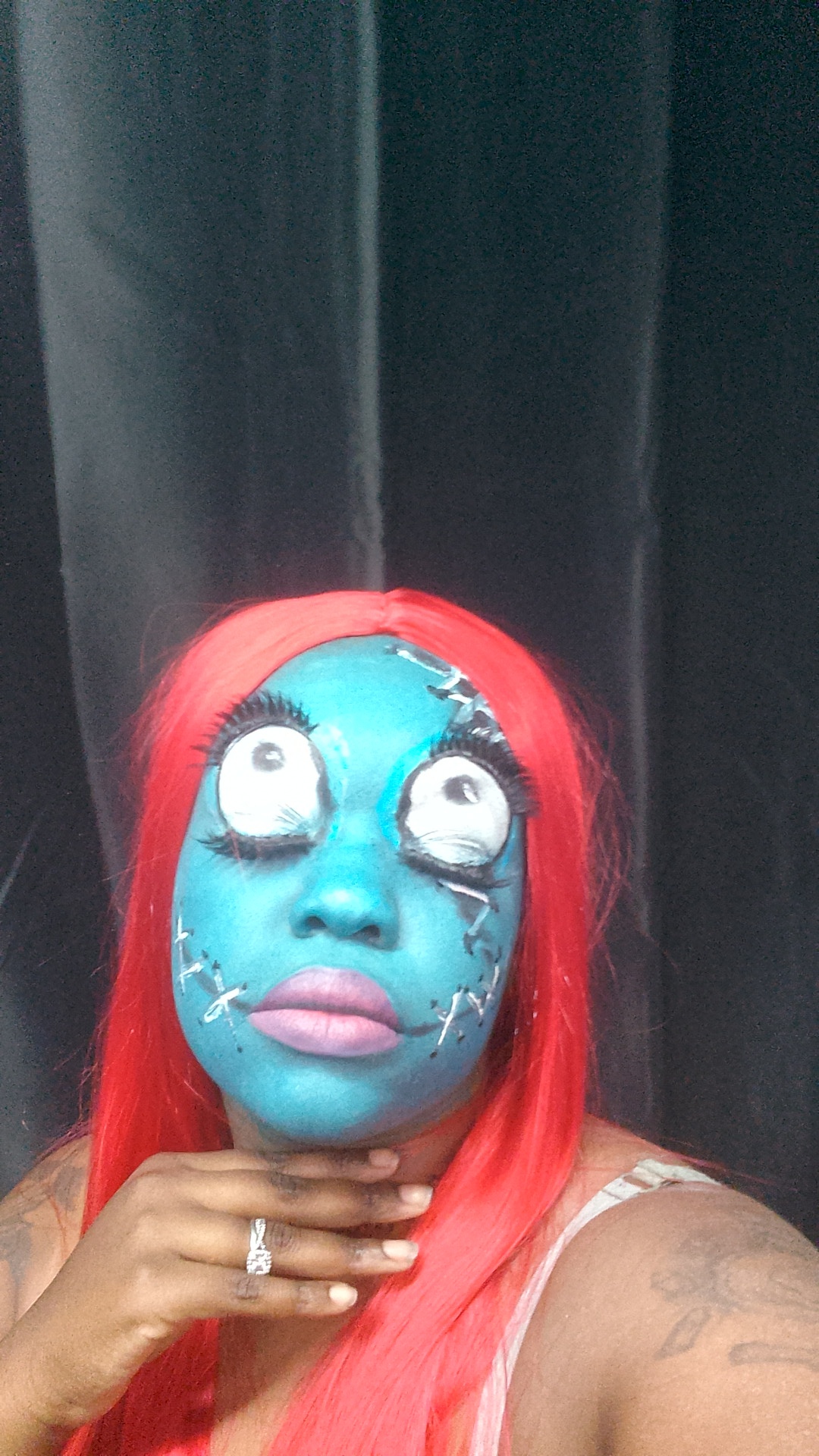 Person with blue face paint, red hair, and large eye makeup, holding chin with hand, black curtain background.