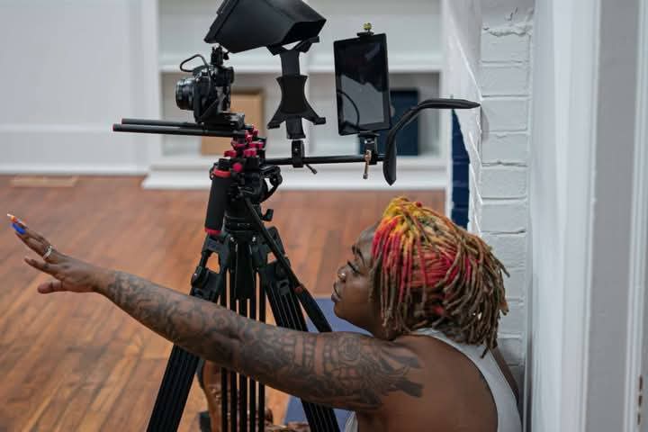 Person with colorful dreadlocks reaching towards a camera mounted on a tripod indoors.