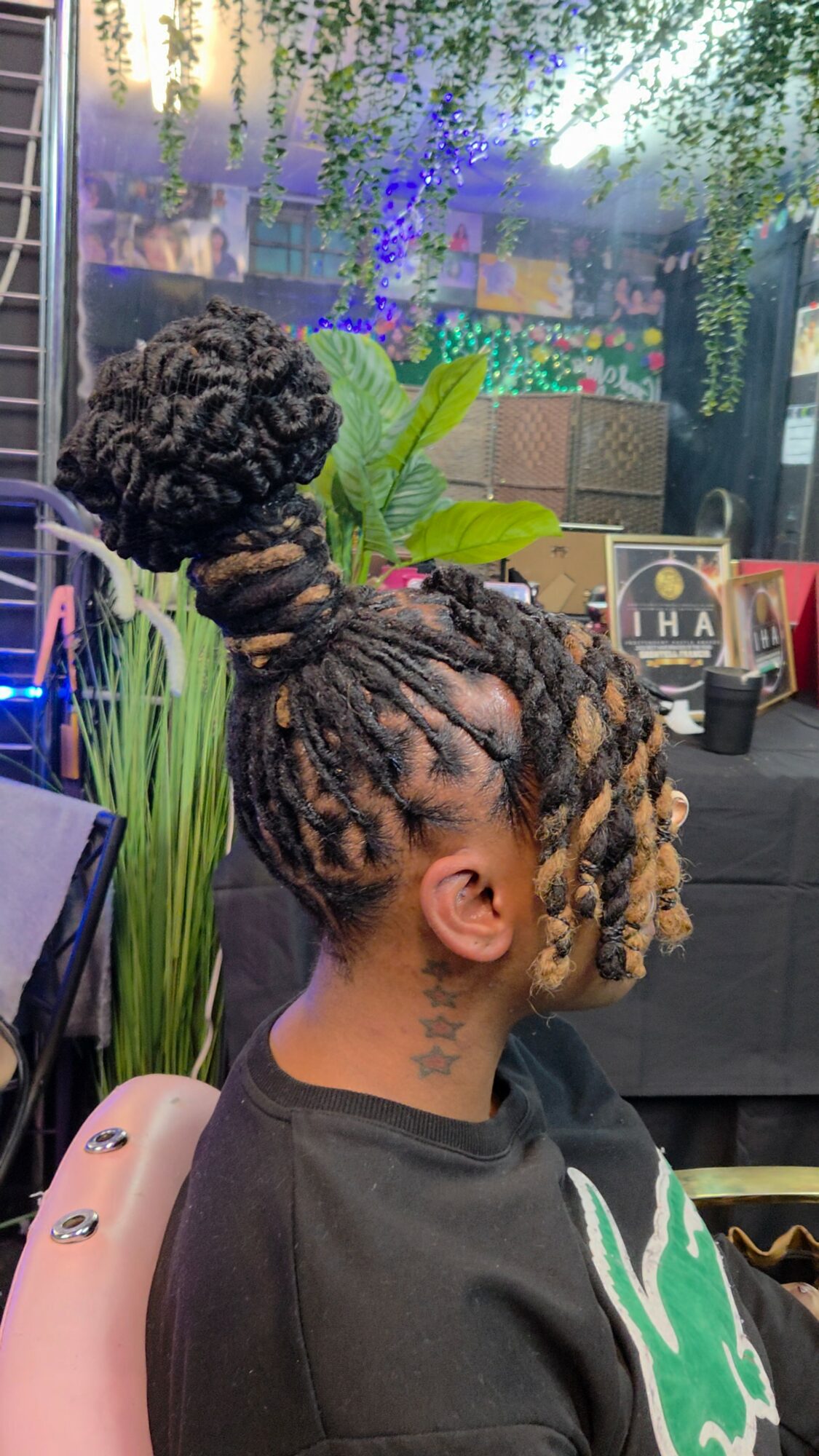 Side view of a person with styled dreadlocks in a high bun, sitting indoors near a window with plants.