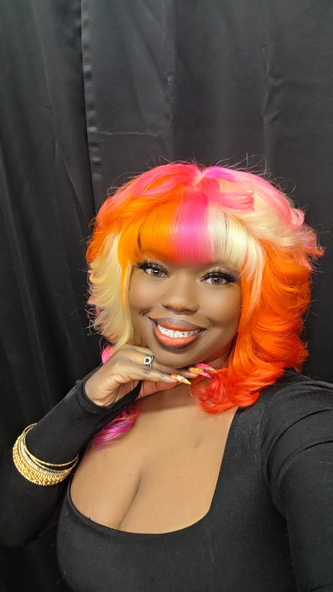 Woman with multicolored hair smiling, black top, black background, hand near face, jewelry on wrist.
