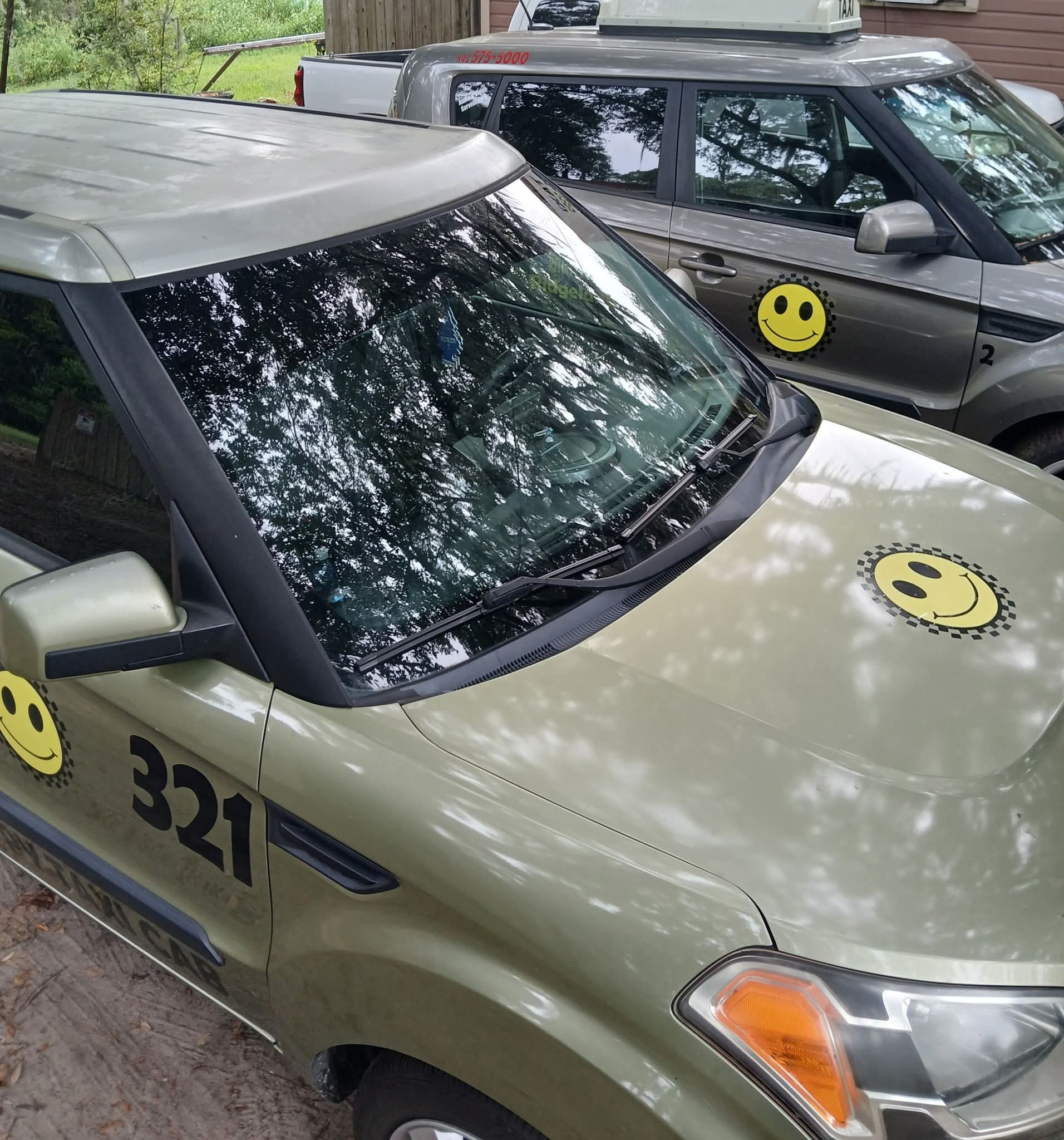 Two cars parked outdoors with smiley face stickers on the hoods and side mirrors, trees reflected on the windshields.