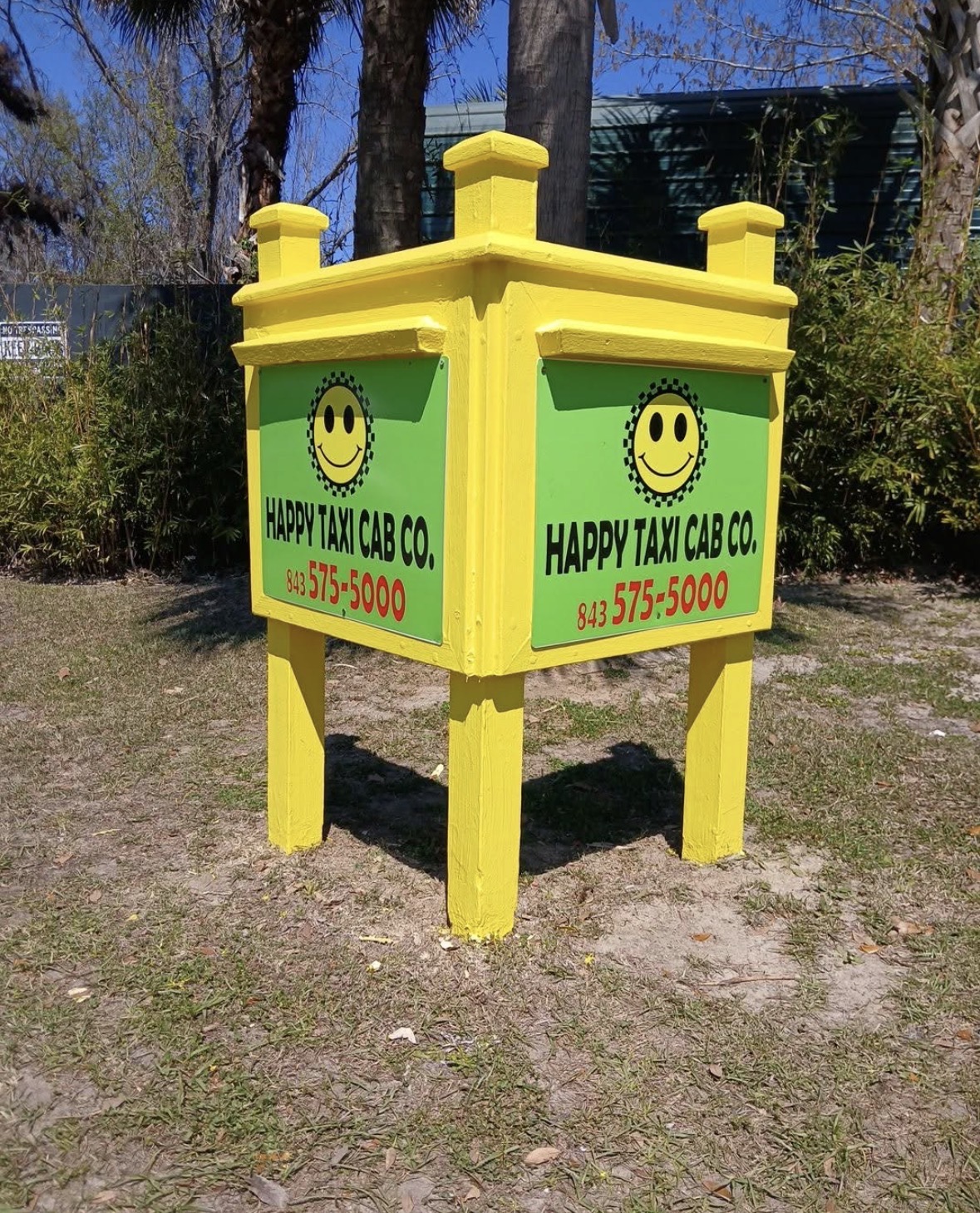 Bright yellow taxi cab sign with green sides, smiley face logo, and contact number, standing outdoors on grass.