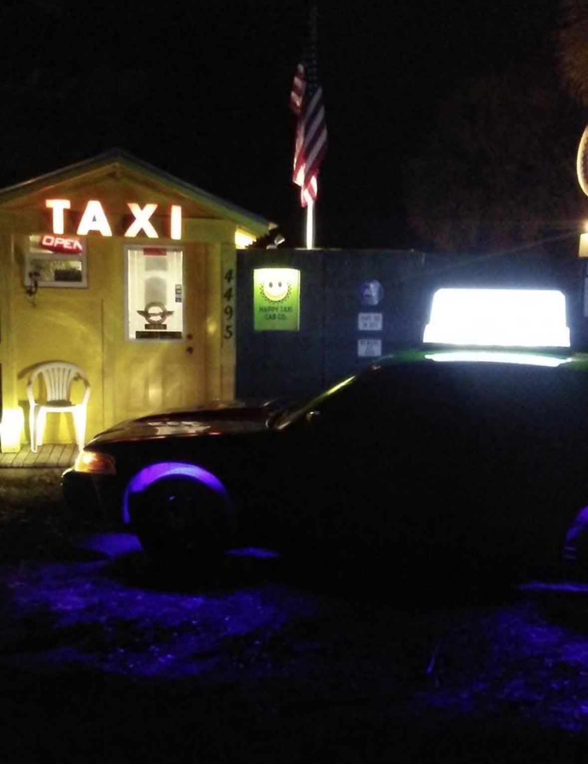 Taxi stand at night with illuminated sign, yellow building, and a dark vehicle in foreground.