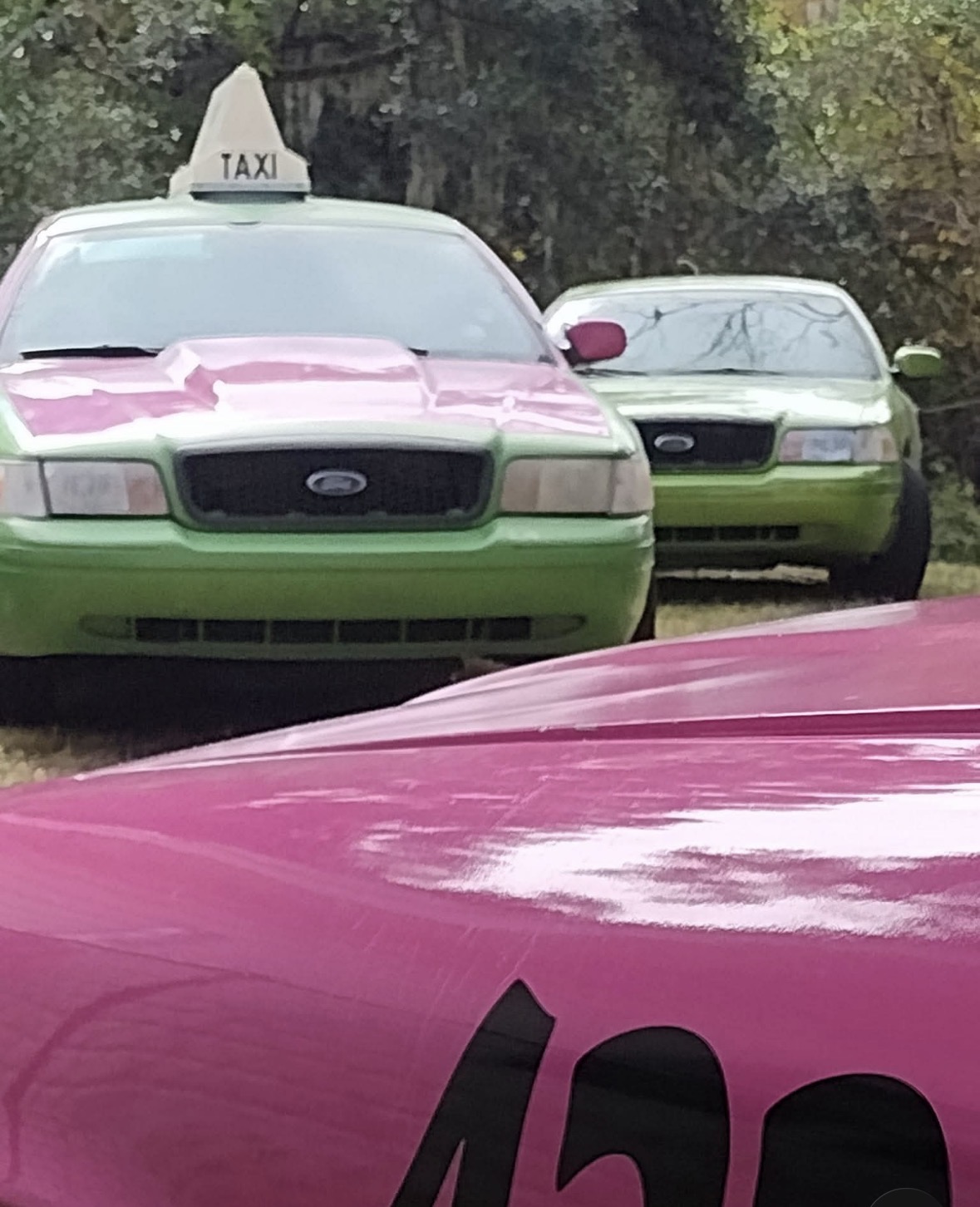 Two cars parked outdoors, one with a taxi sign on top, and a pink vehicle in the foreground.