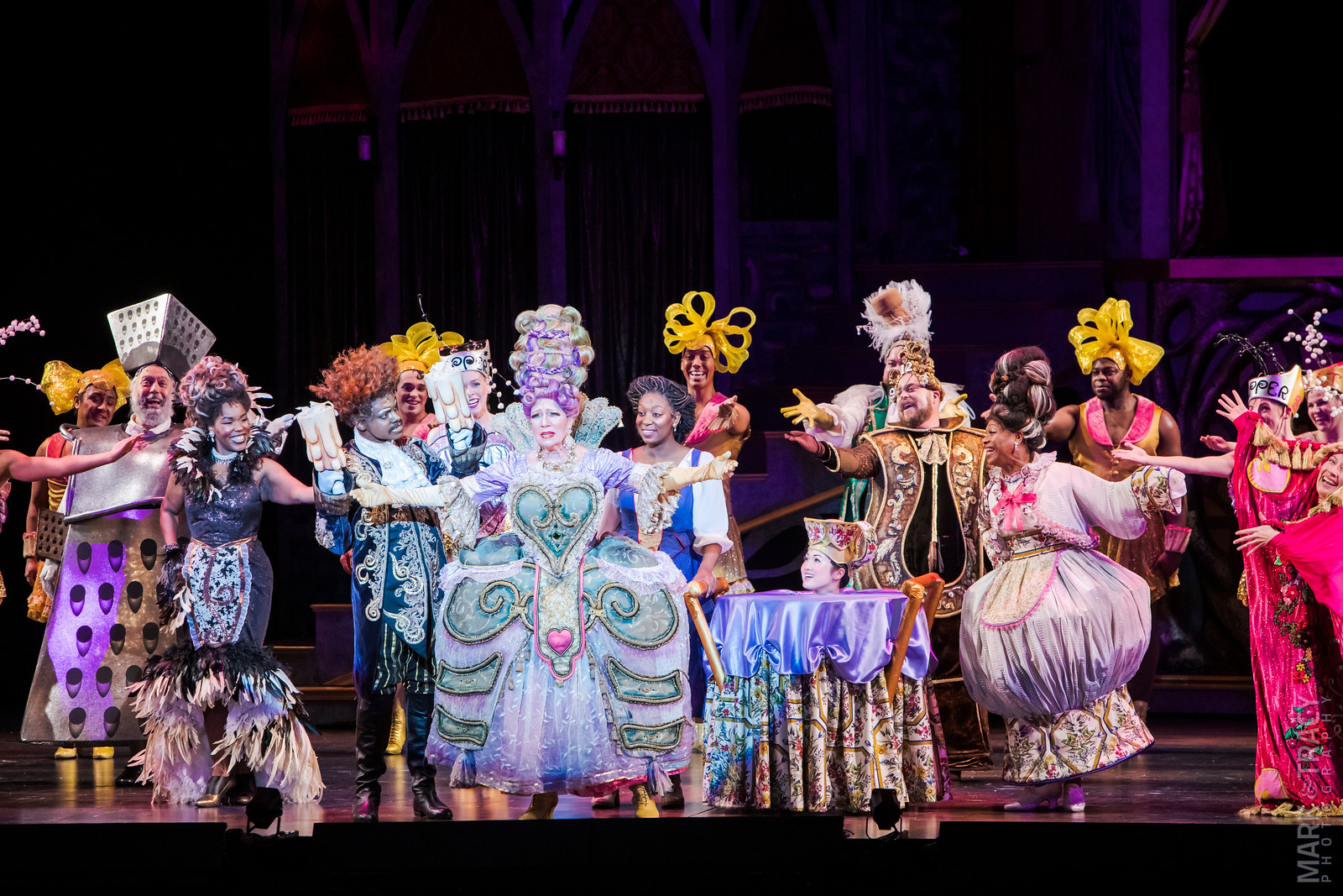 Group of performers in colorful costumes on stage, some with masks and props, in a theatrical scene.