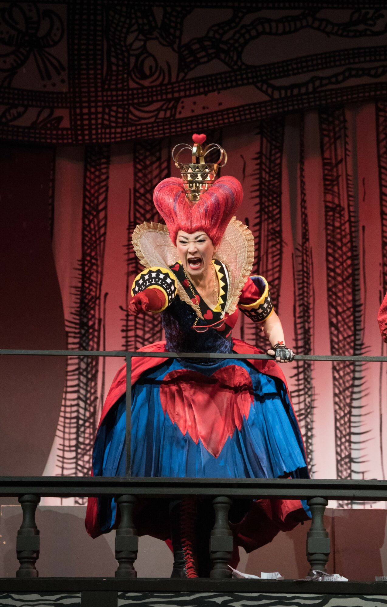 Performer in colorful costume on stage, with a red and blue skirt and elaborate headpiece, behind a barrier.