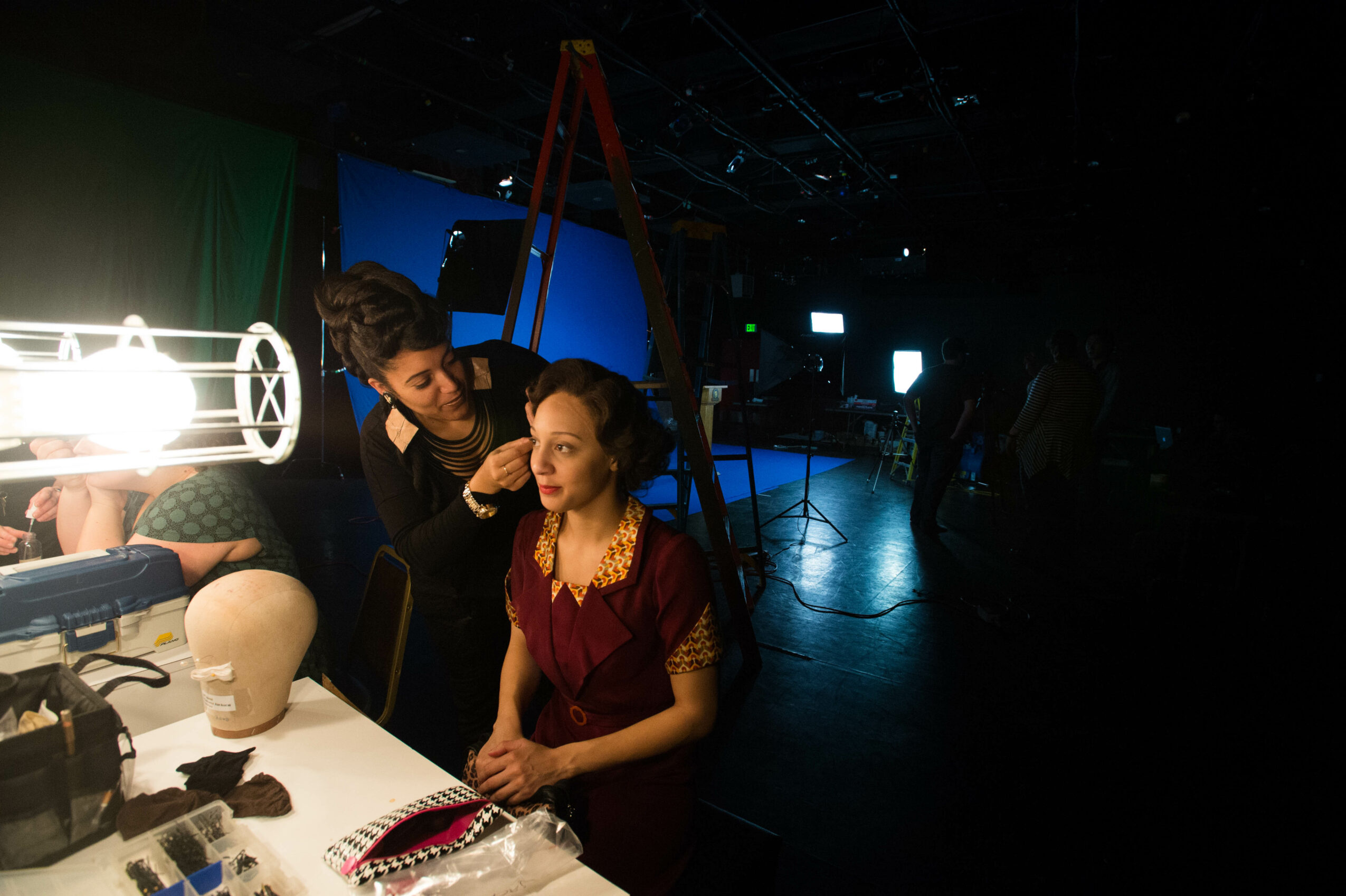Woman sitting at a table with makeup artist applying makeup, ladder in background, dark studio setting.