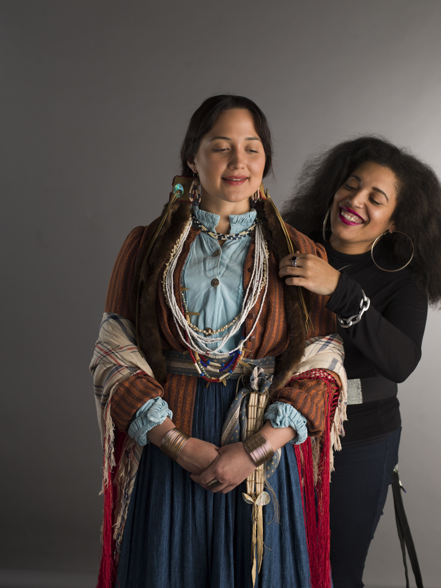 Two women smiling, one with long hair and colorful jewelry, the other with curly hair and hoop earrings, standing close together.