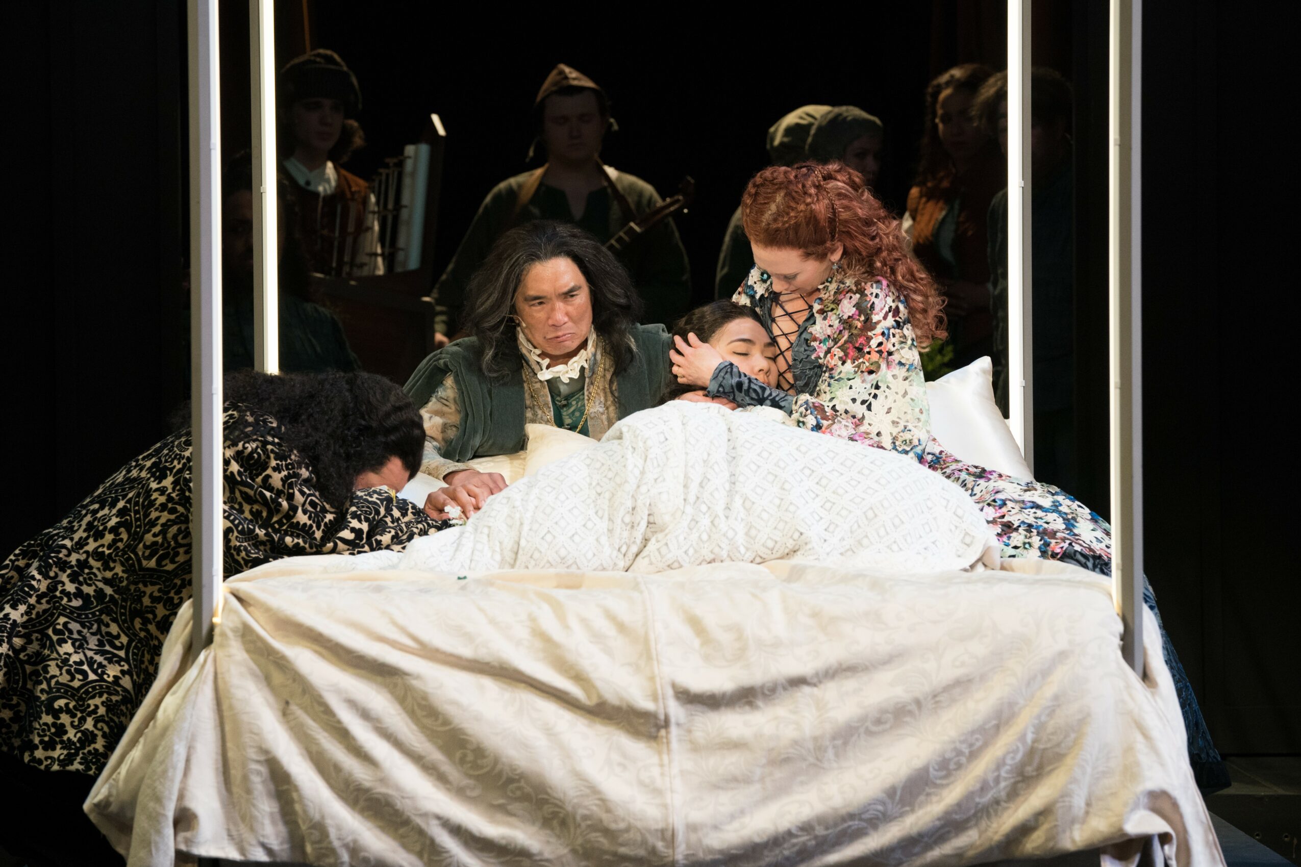 Group of women gathered around a bed with a person lying on it, in a dimly lit room.