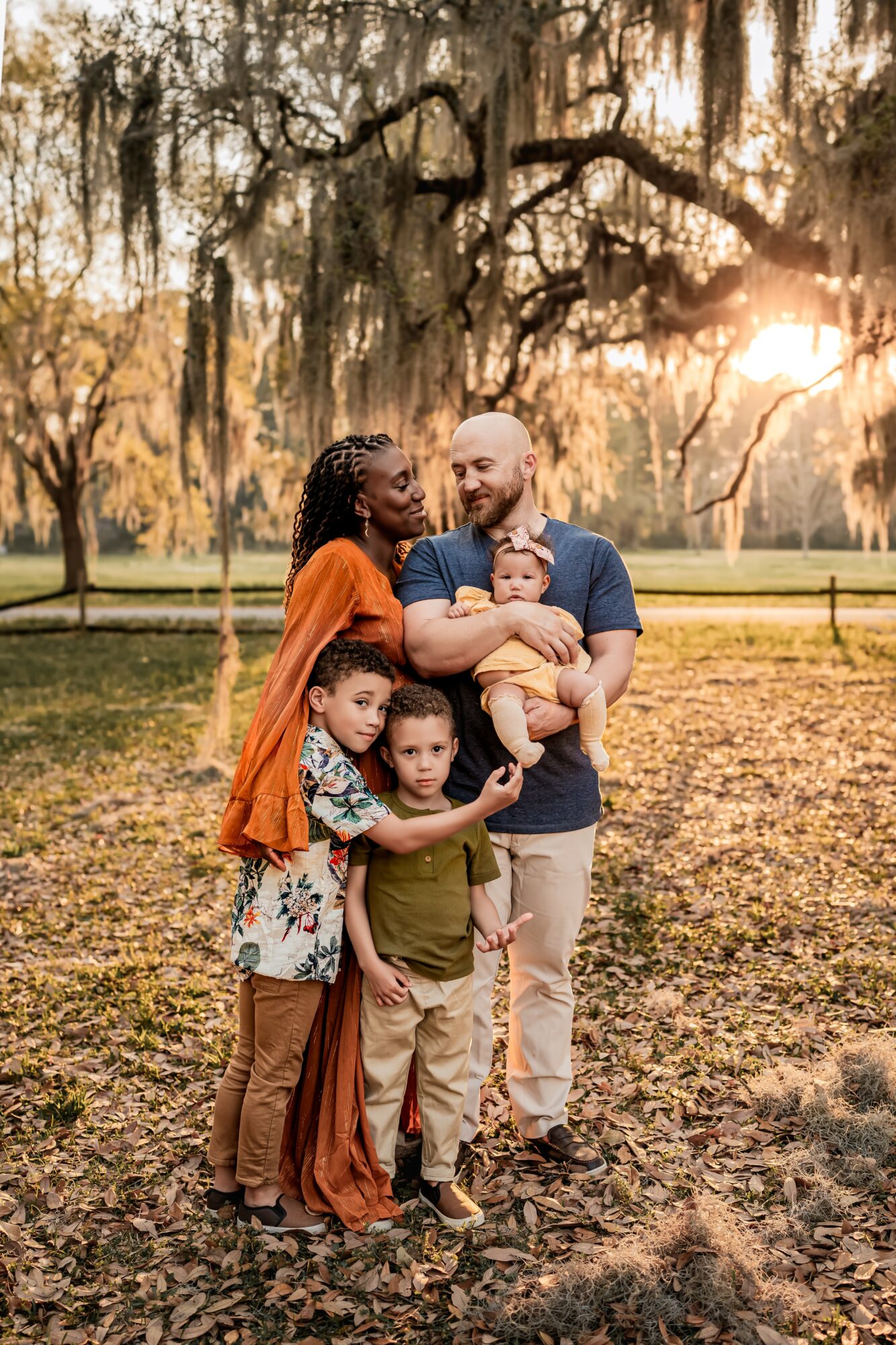 Family of five outdoors in a park with trees and sunlight, smiling and holding children, autumn leaves on ground.