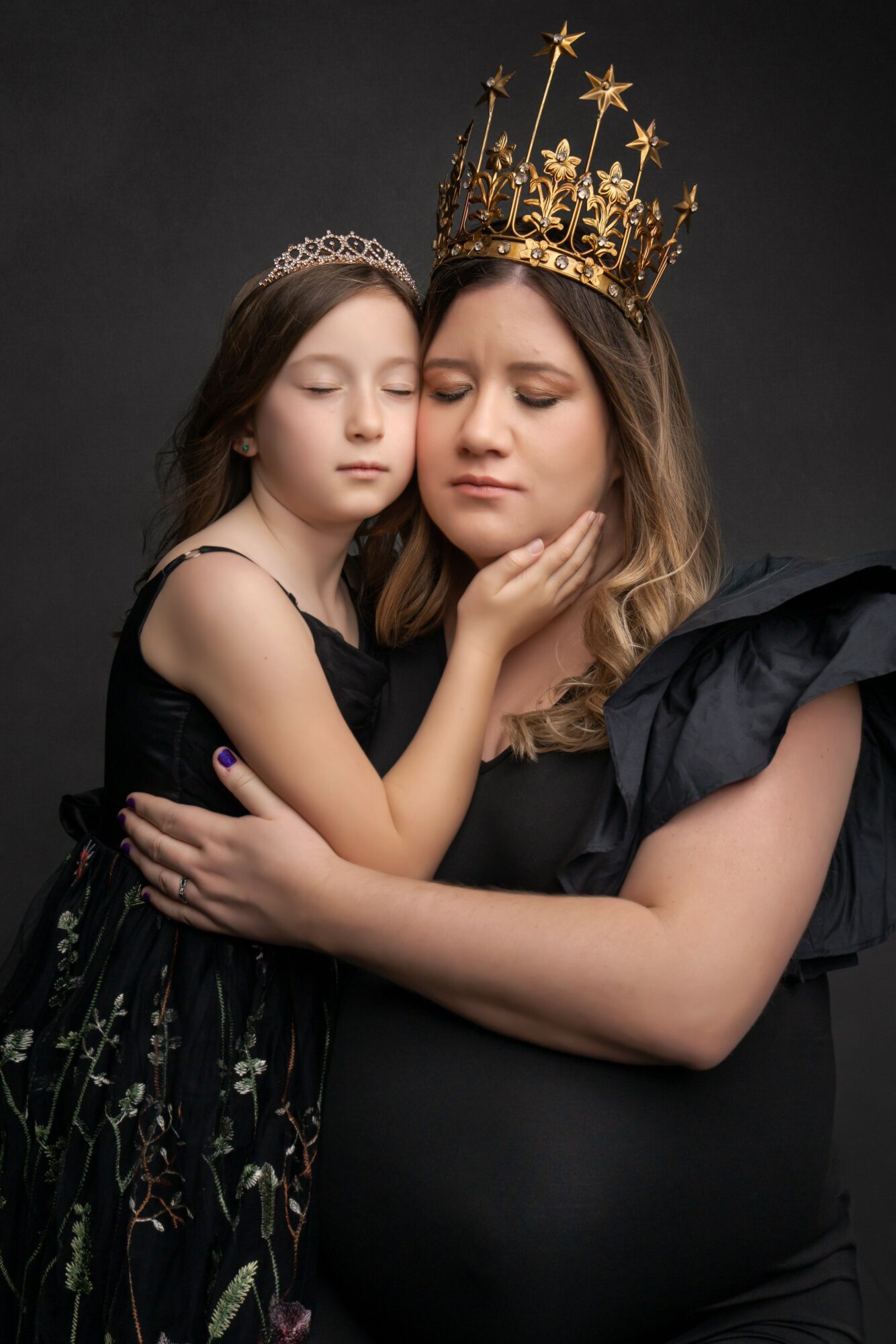 Young girl and woman with crown, close embrace, dark background, girl touches woman's face, both with closed eyes.