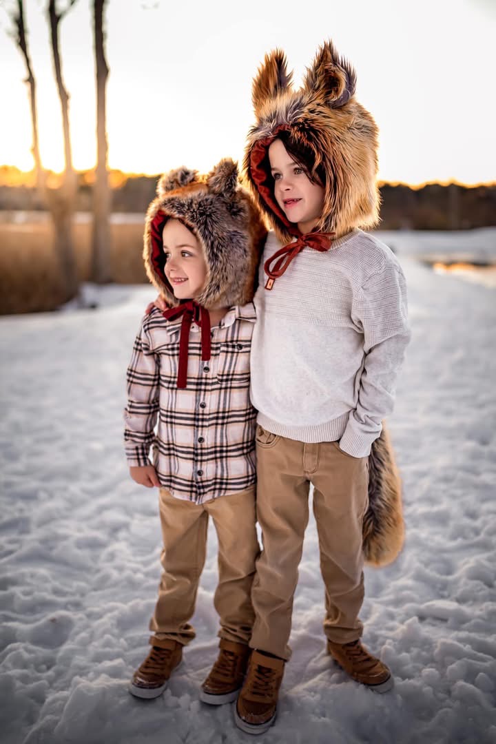 Two children wearing animal hats stand on snow-covered ground during sunset, with trees in the background.