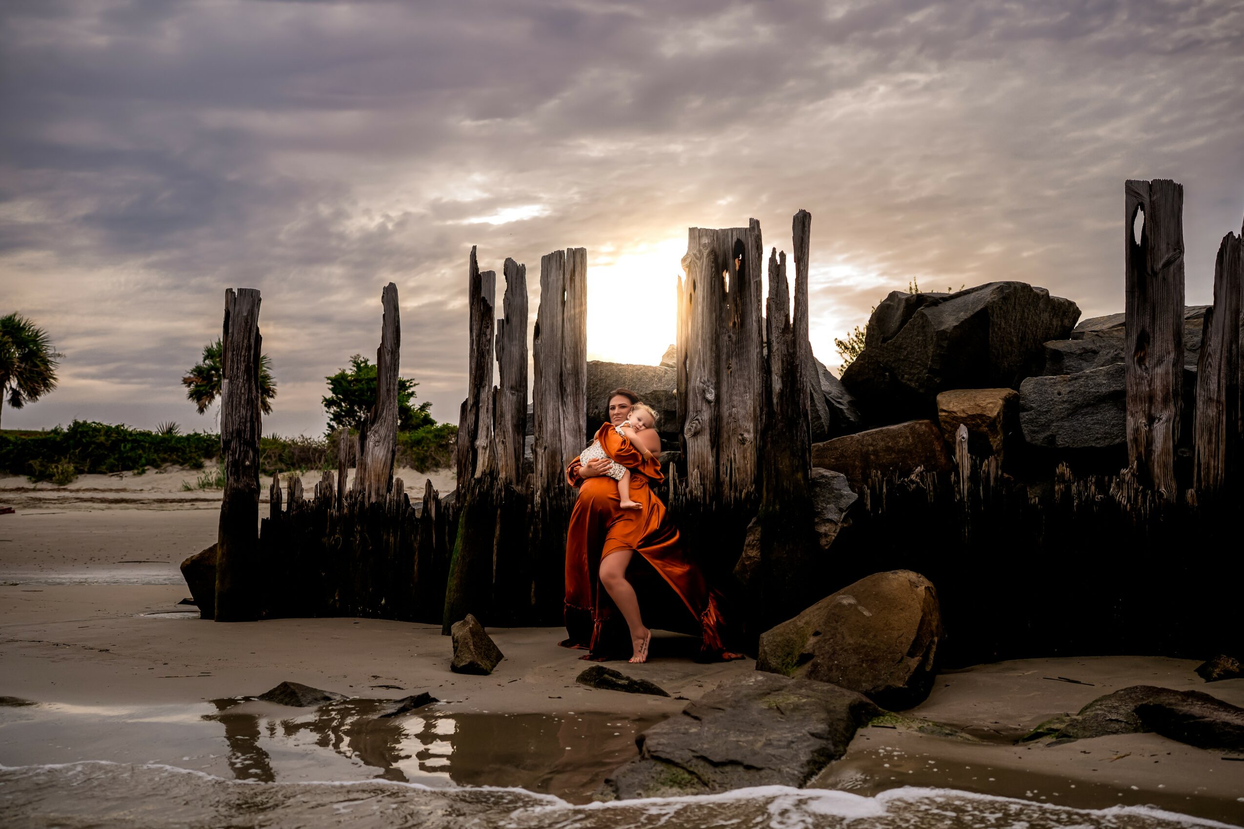 Person standing on a beach near weathered wooden posts and rocks with cloudy sky and sunset in background.