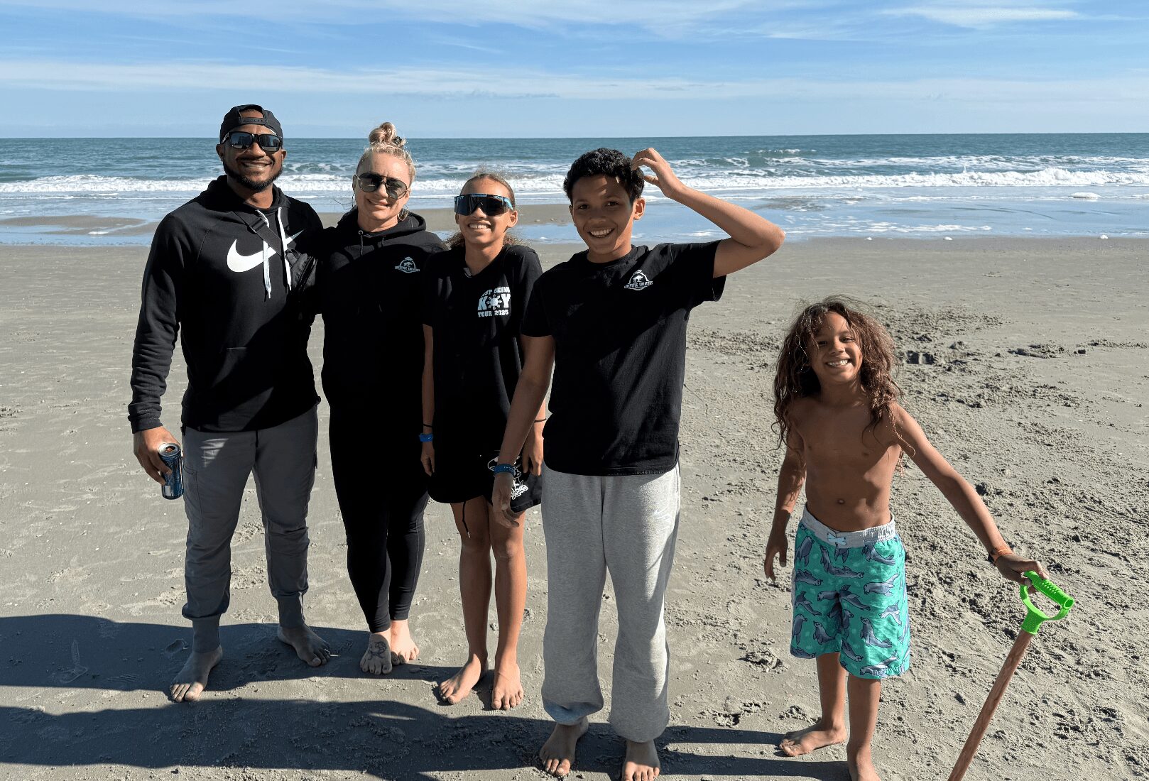 Five people standing on a beach with ocean waves in the background, smiling and posing for the photo.