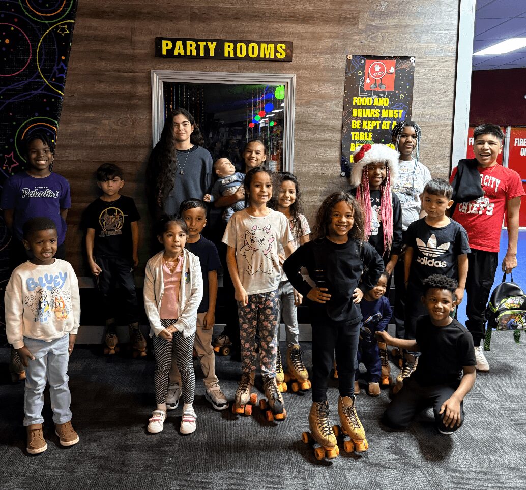 Group of children and adults posing in front of a party rooms sign and colorful wall, some smiling and standing.