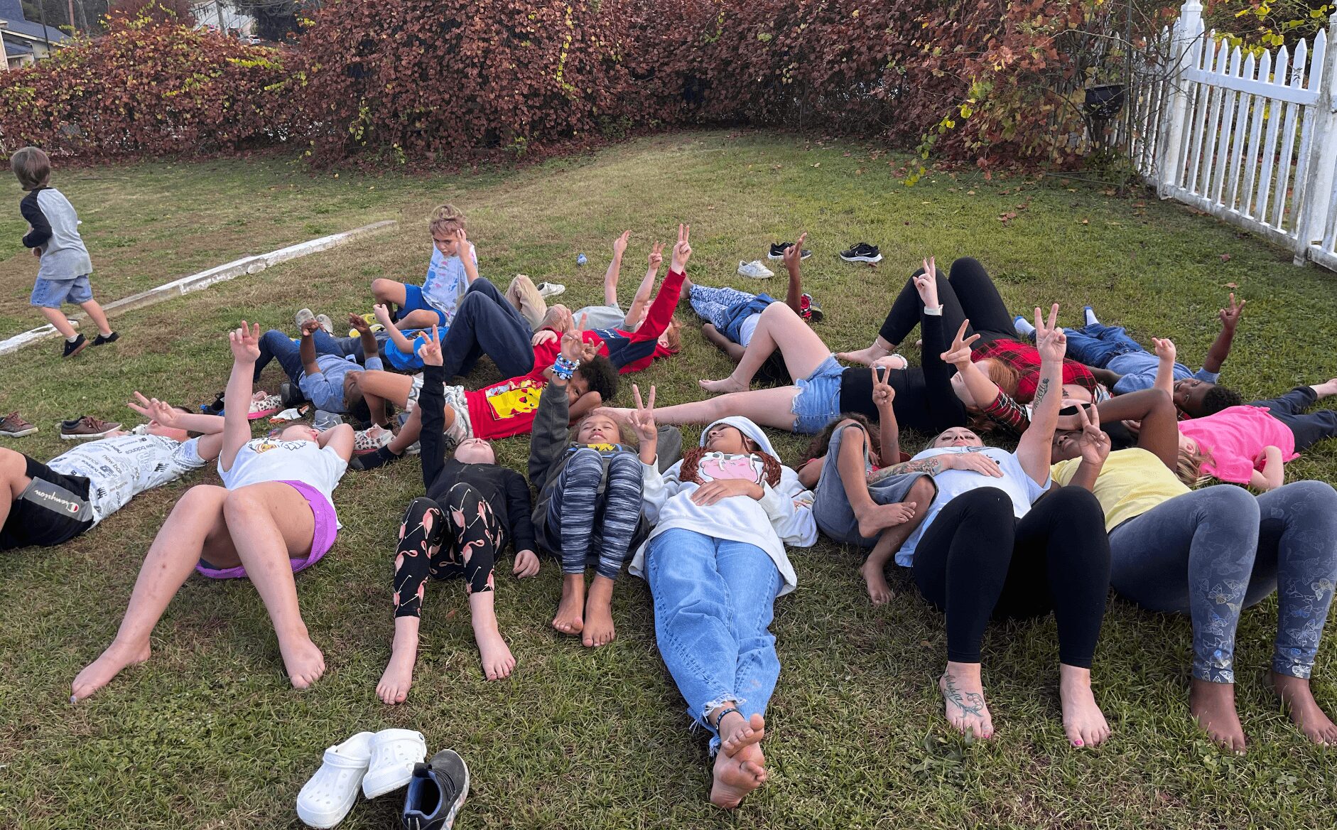 Group of children lying on grass with arms raised, outdoors in a yard with a white fence and bushes.