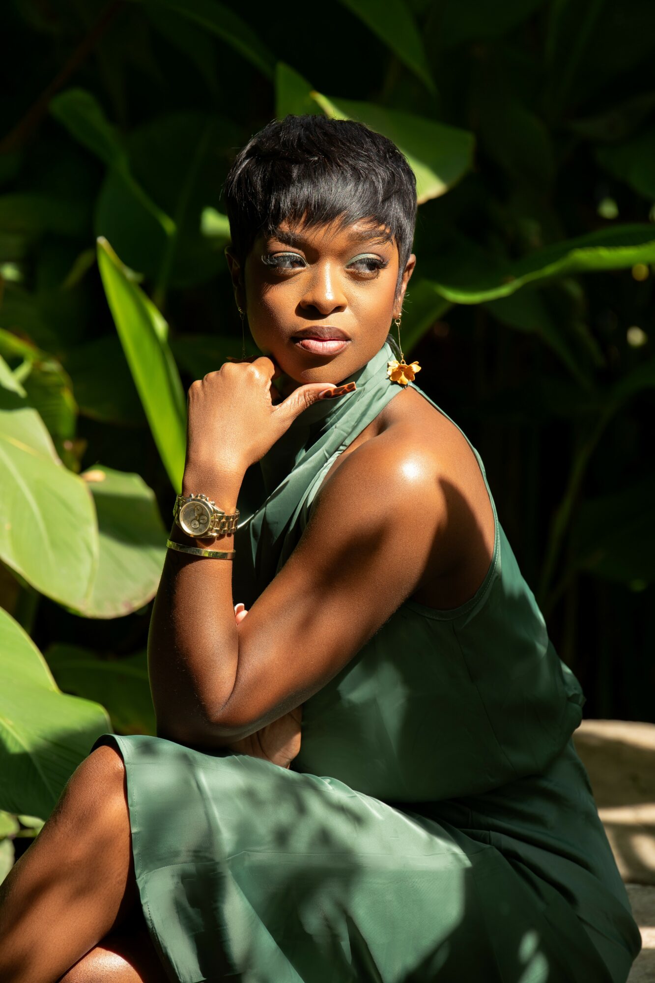 Woman with short hair and a green dress sitting among large green leaves, looking to the side.