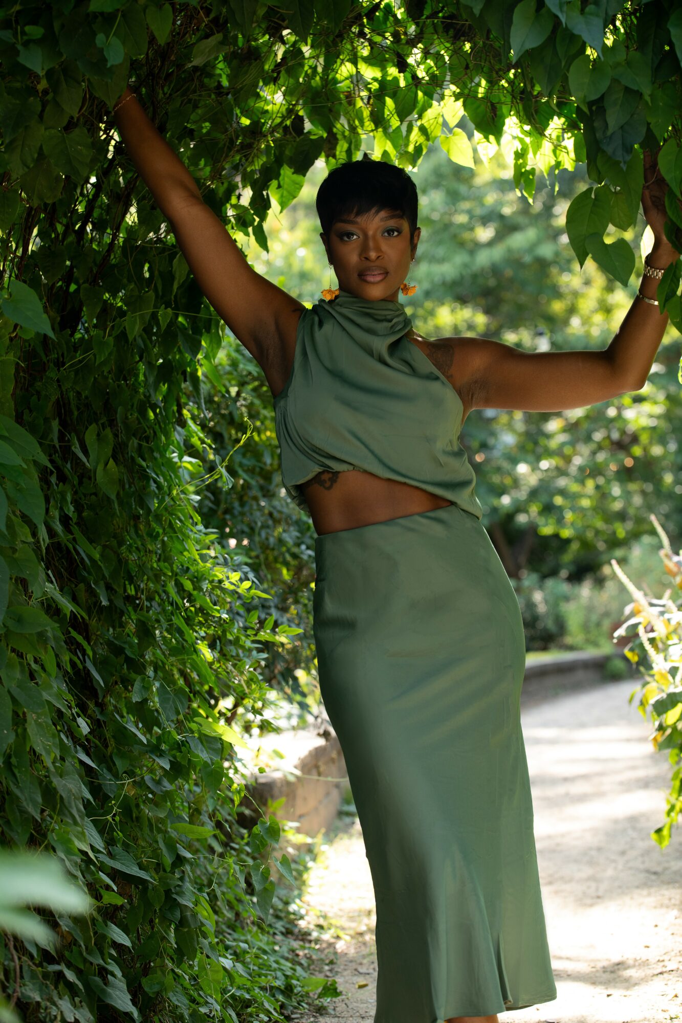 Person standing outdoors among green foliage, wearing a green outfit, with arms raised and smiling.