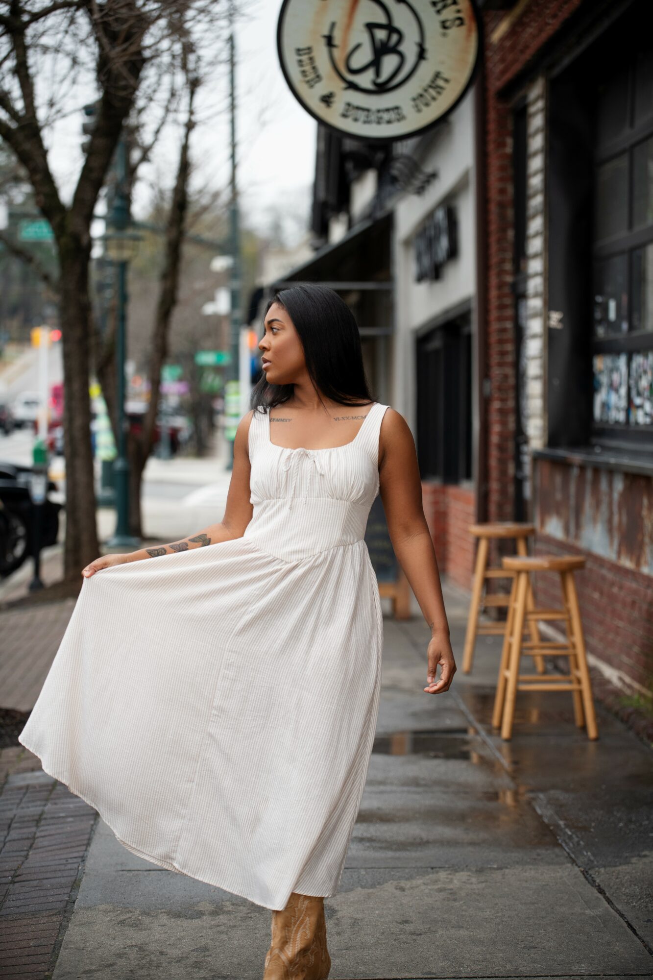 Woman in white dress walking on sidewalk near storefronts and trees, holding her dress.