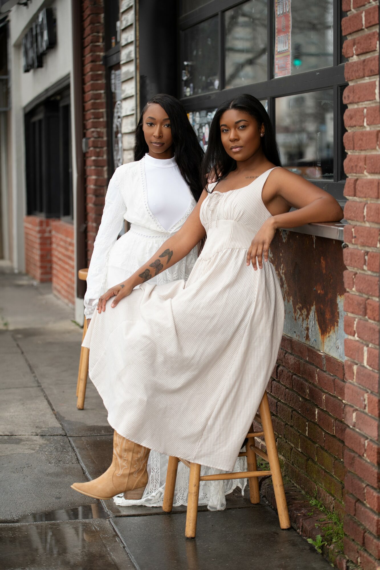 Two women pose outdoors against a brick wall, one sitting on a stool and the other standing beside her.