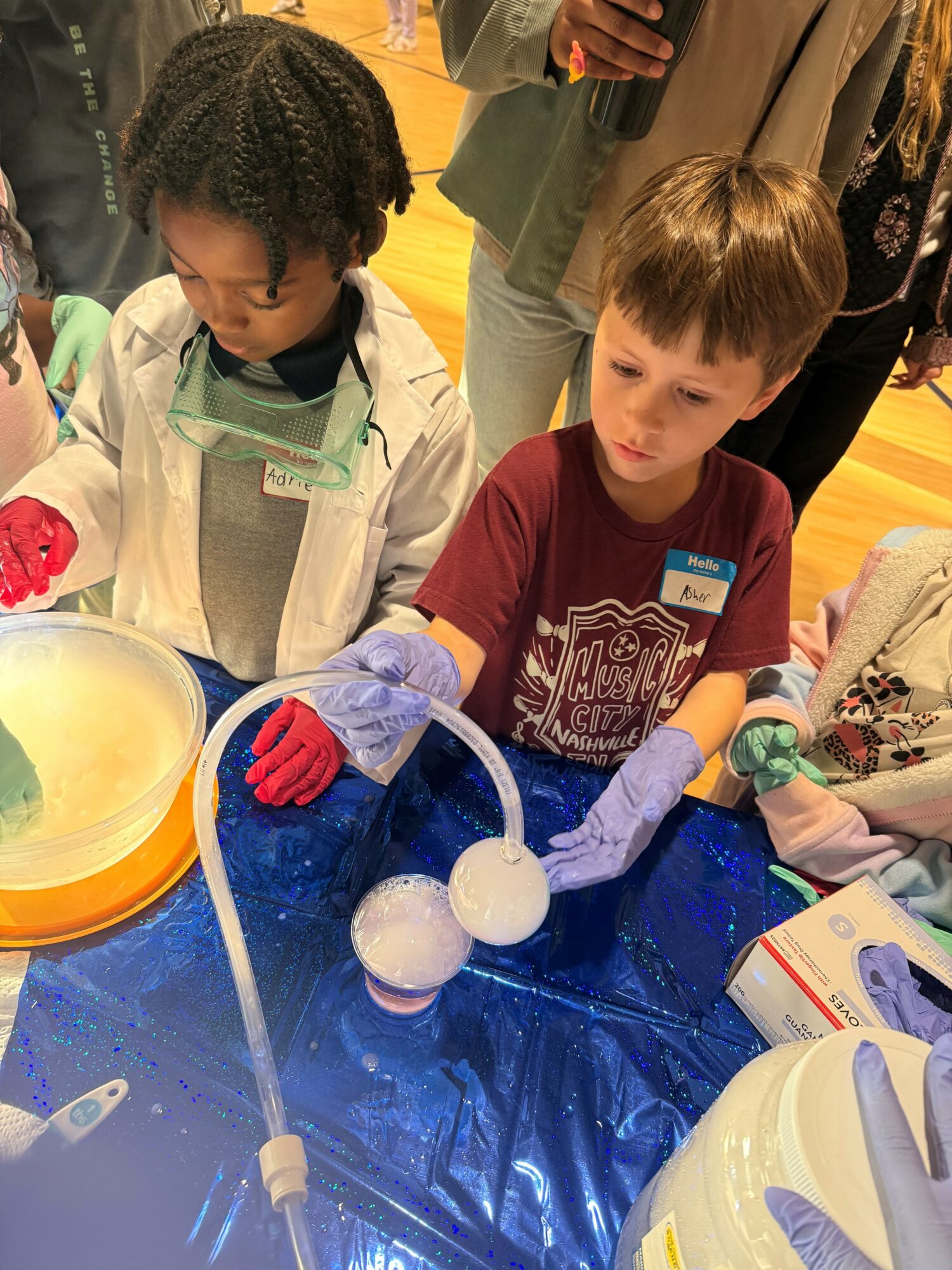 Two children conduct a science experiment with balloons and liquids at a table, surrounded by adults in an indoor setting.