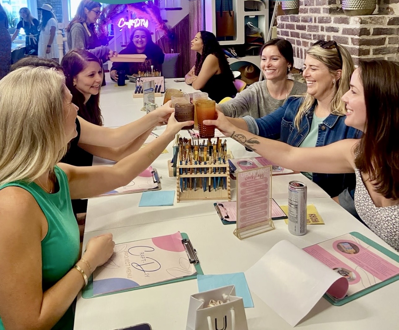 Group of women at a table raising drinks in a toast, smiling, in a lively restaurant or bar setting.