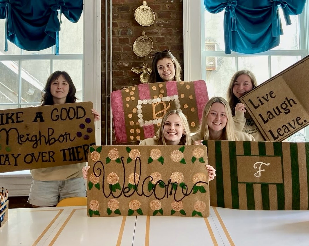 Five smiling women holding decorated signs in a room with windows and blue curtains.