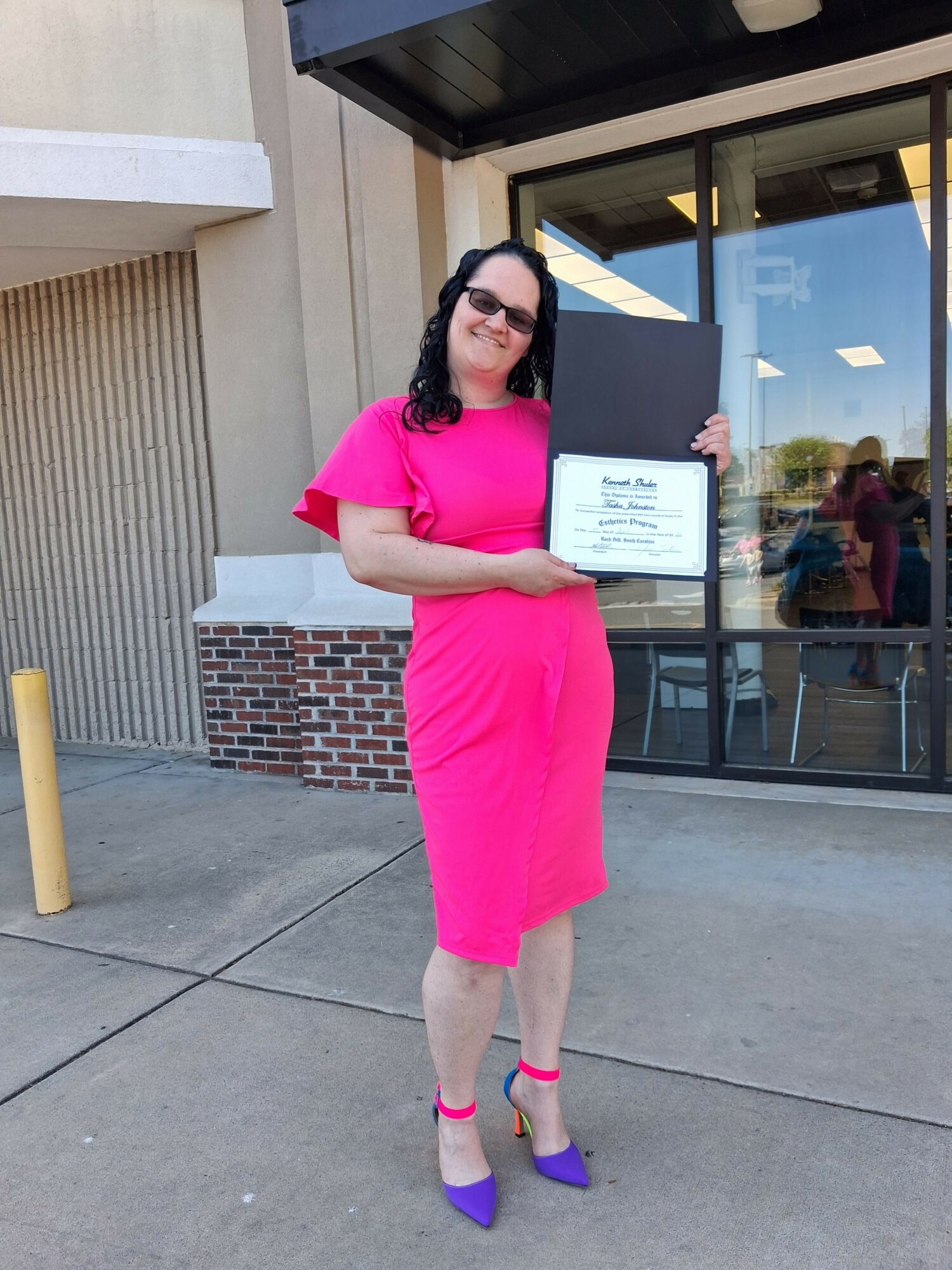 Woman in pink dress holding a certificate outside a building with glass windows.