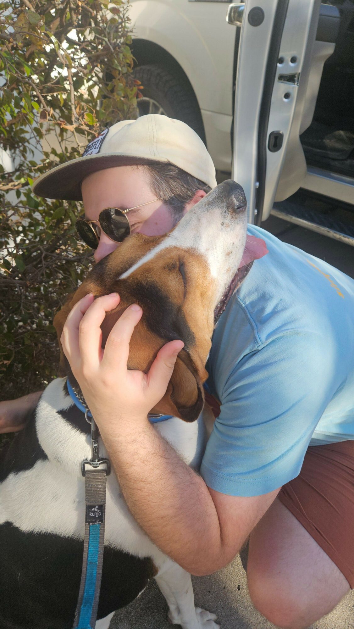 Person hugging a dog outdoors, wearing sunglasses, hat, and blue shirt.